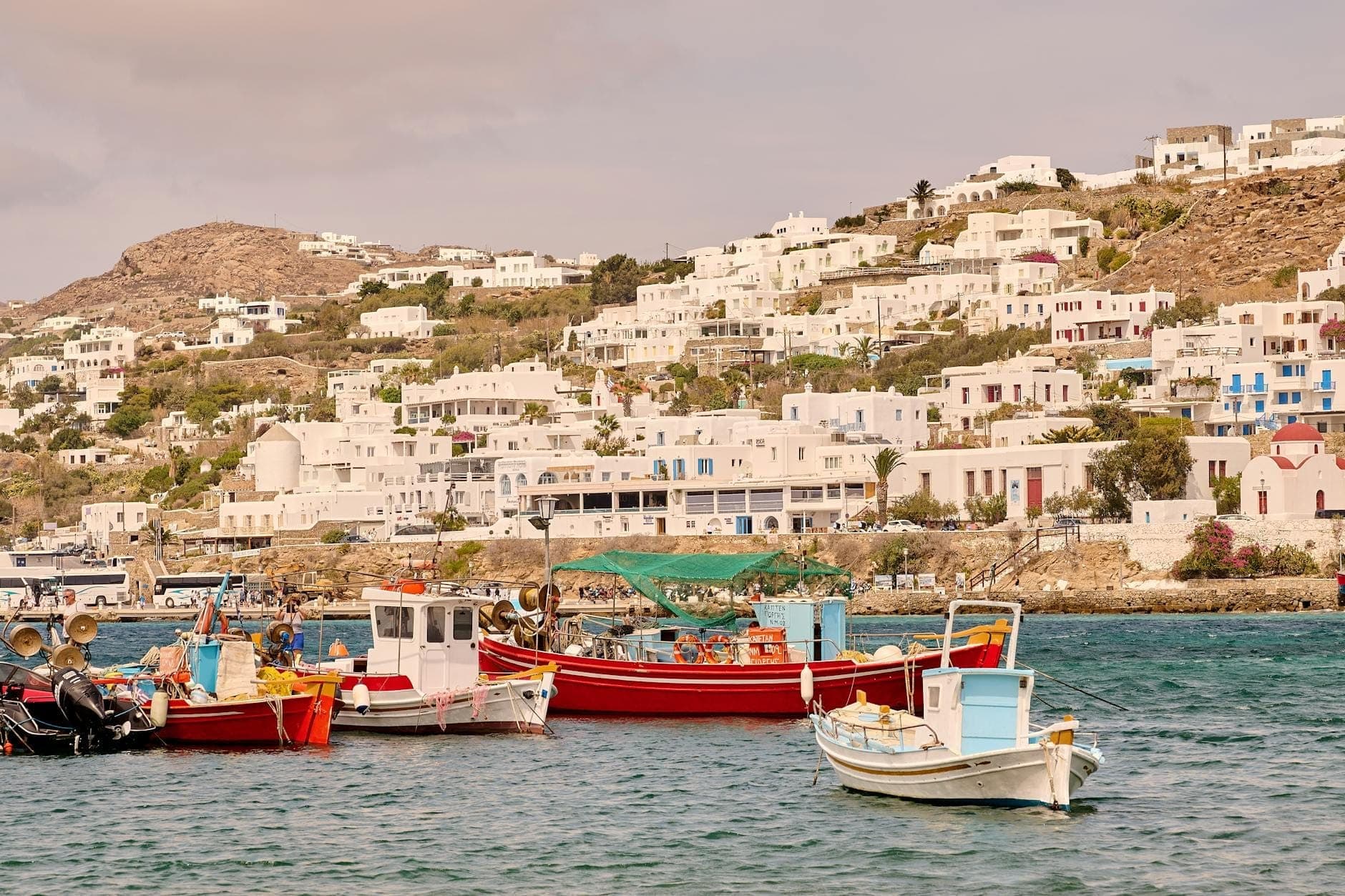 Traditionelle Fischerboote in türkisfarbenem Wasser vertäut, mit den weiß getünchten Häusern von Mykonos im Hintergrund auf dem Hügel.
