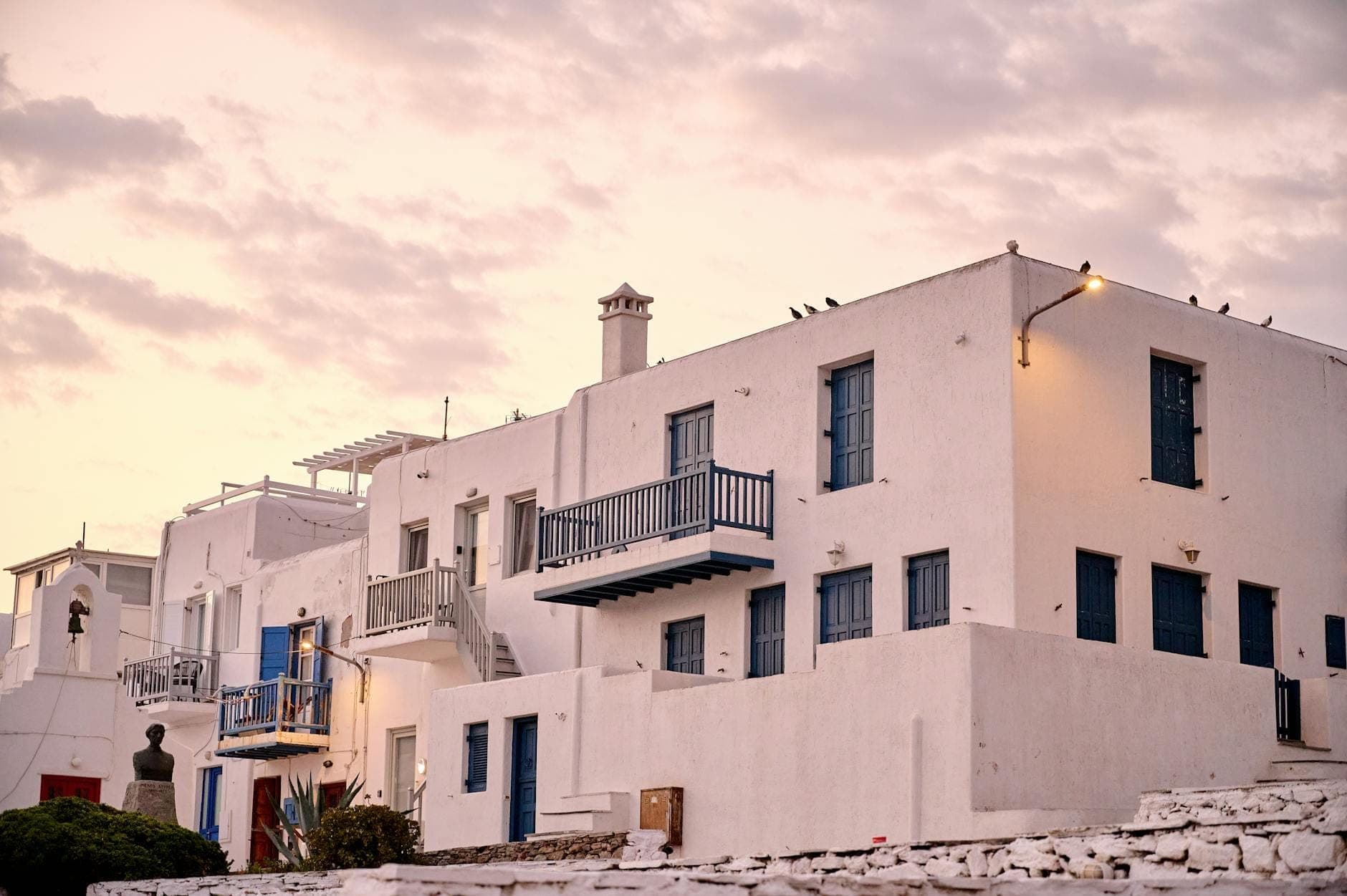 Weiß getünchte Gebäude mit blauen Balkonen und Fenstern in Mykonos bei Sonnenuntergang, typisch für die lokalen Unterkunftsmöglichkeiten.