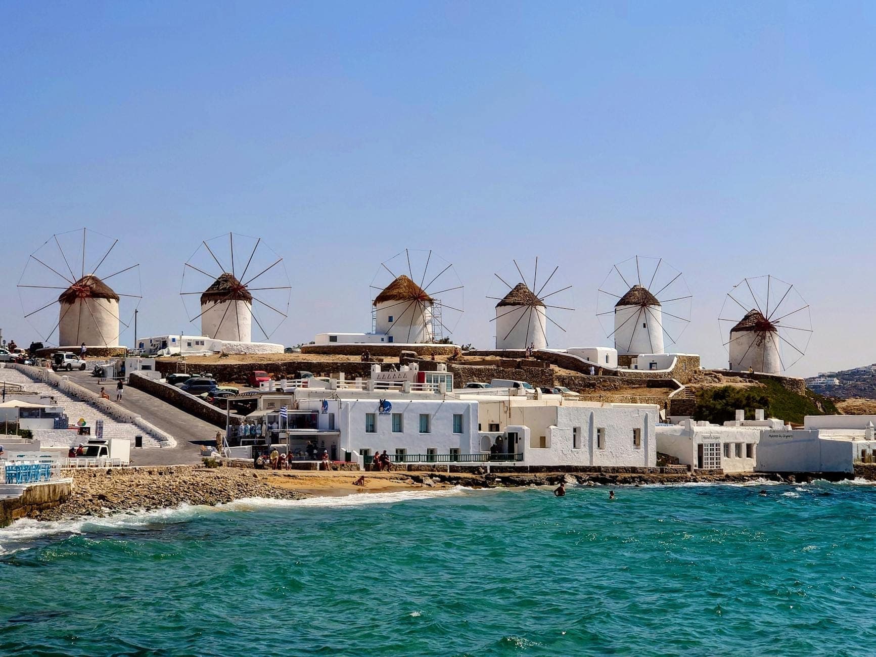 Row of iconic Mykonos windmills on a hill overlooking the sea, with whitewashed buildings and turquoise waters under a clear blue sky.