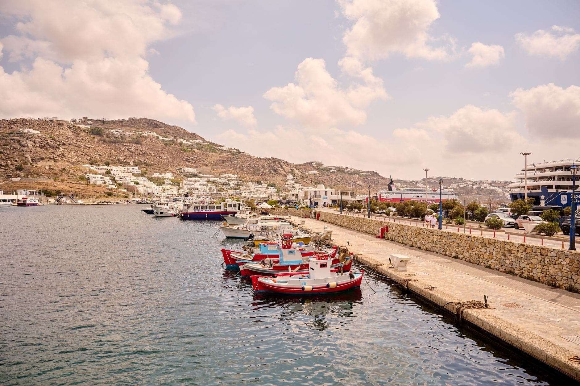 Mehrere Boote, die am Kai des neuen Hafens von Mykonos festgemacht sind, mit Stadtgebäuden und Hügeln im Hintergrund.