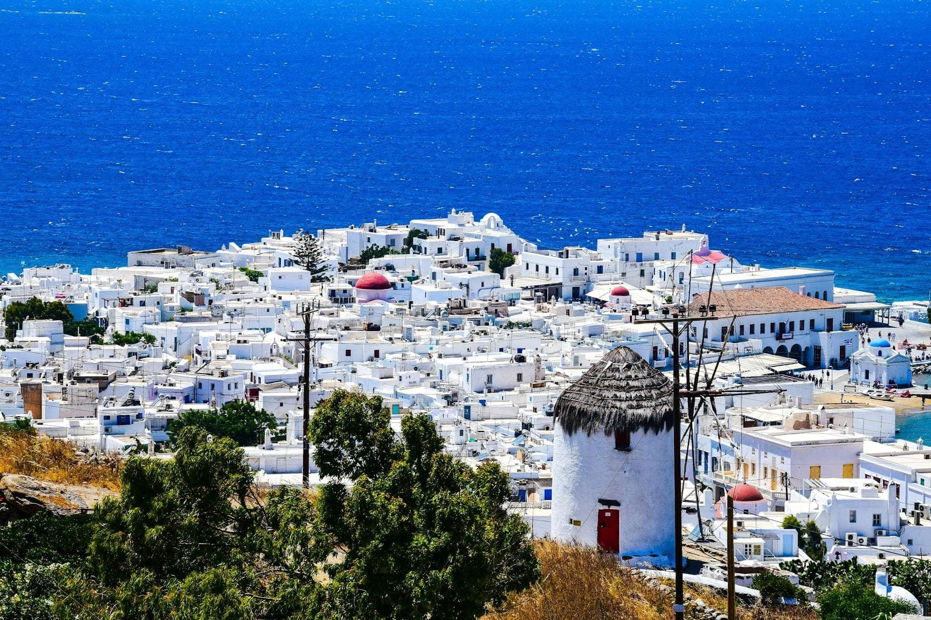 Panoramablick auf Mykonos Town mit weißen kykladischen Häusern, einer Windmühle und dem tiefblauen Meer im Hintergrund unter einem klaren Himmel.