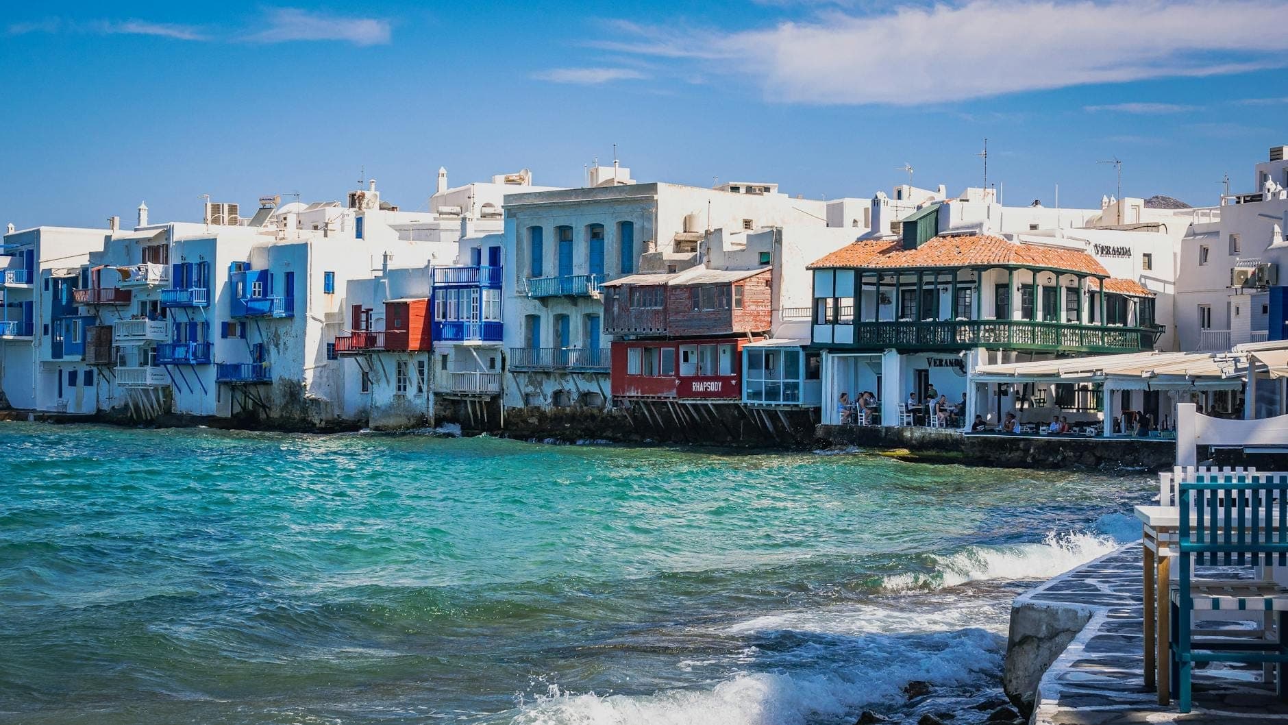 Lebhafte Bars und Restaurants am Wasser in Little Venice auf Mykonos, mit plätschernden Wellen und entspannten Gästen an Außentischen.