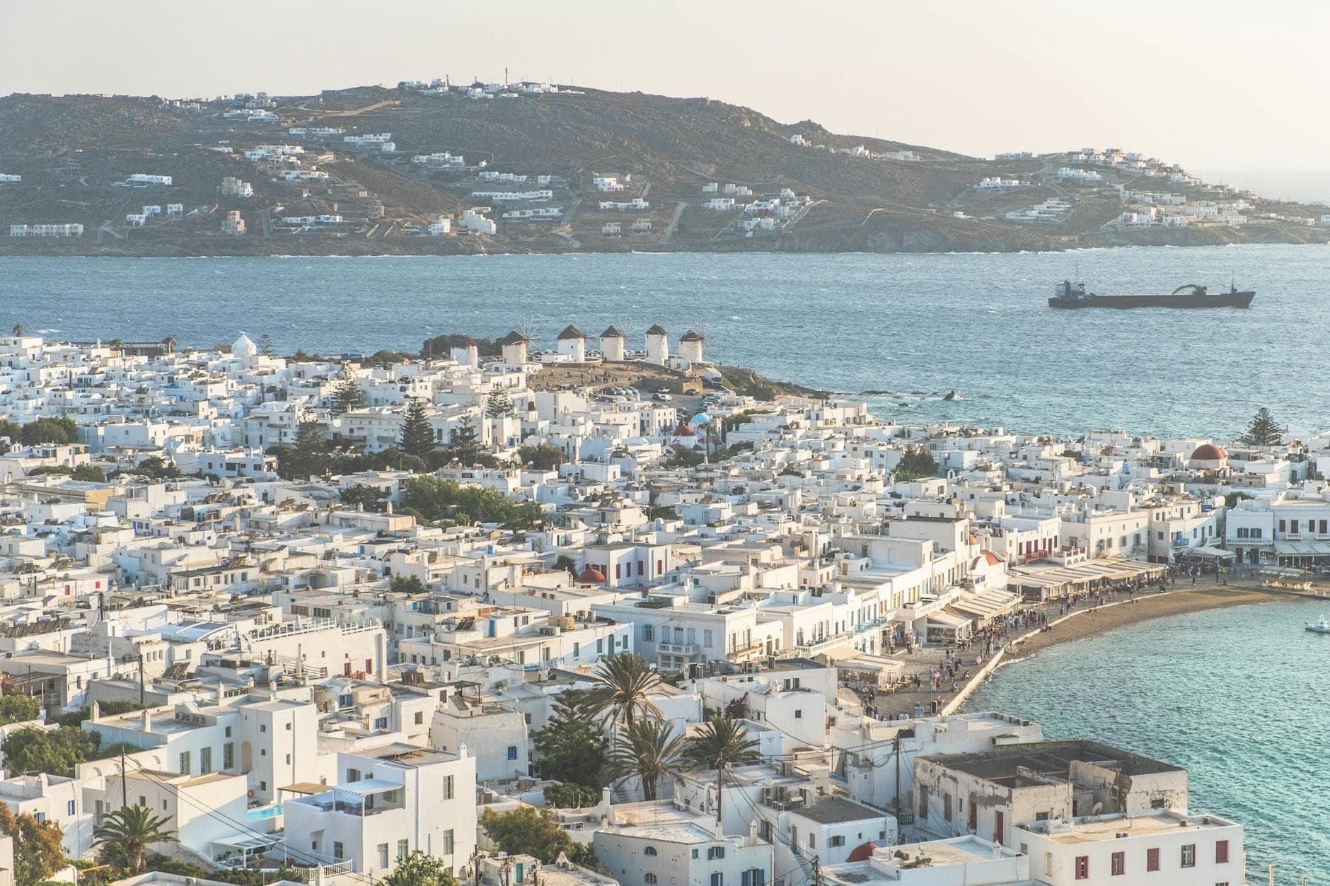 Weitläufige Luftaufnahme der Stadt Mykonos mit weiß getünchten Gebäuden, Küstenlinie, Hafen und ikonischen Windmühlen, mit Hügeln und Meer im Hintergrund.