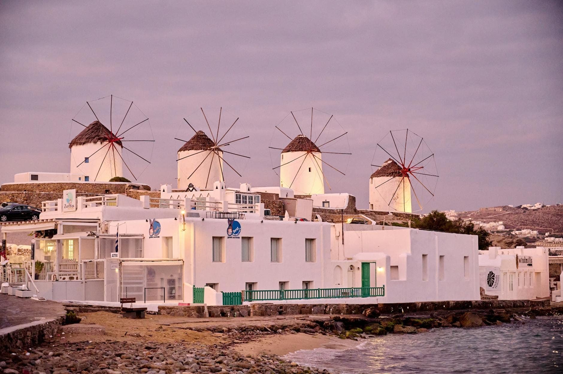 Vier ikonische weiß getünchte Windmühlen mit Reetdächern stehen auf einem Hügel über Mykonos Town, mit Blick auf weiße Häuser und die Ägäis bei Sonnenuntergang.