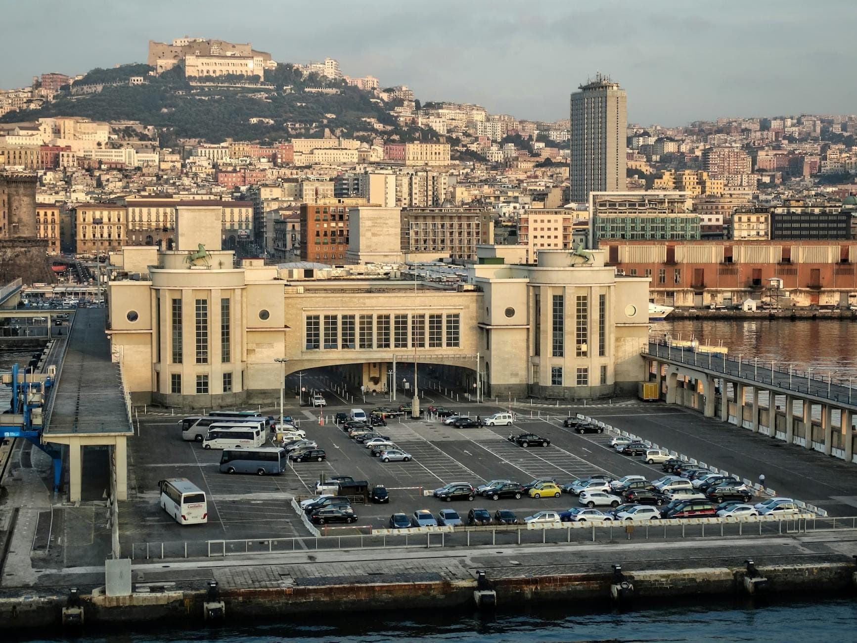 Estacionamento em frente ao terminal do porto de Nápoles com carros, vans e ônibus visíveis, com a paisagem urbana e as colinas de Nápoles ao fundo durante o dia.
