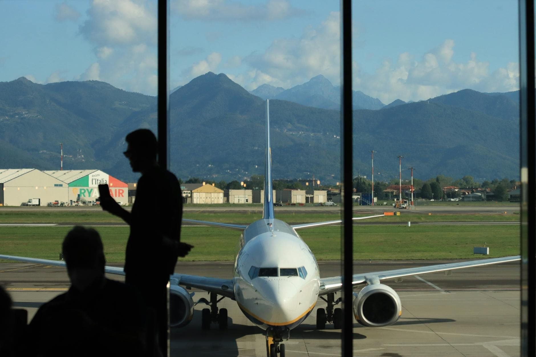 Vista de um avião da Ryanair no Aeroporto de Nápoles (NAP) com pista, edifícios do aeroporto e montanhas verdes ao fundo em um dia ensolarado.