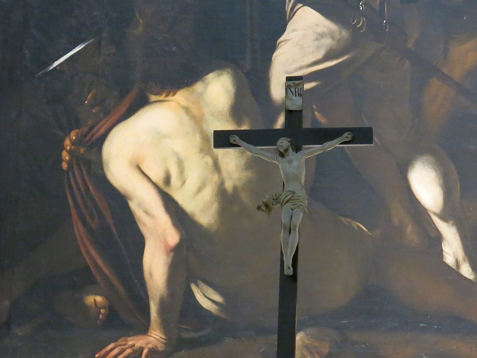 Plano aproximado de um crucifixo em frente à dramática pintura de Caravaggio, com figuras musculosas iluminadas por uma iluminação teatral e intensa.