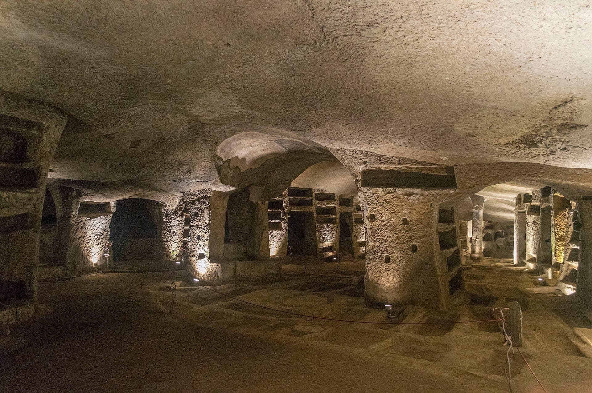 Vista ampla das iluminadas Catacumbas de San Gennaro mostrando pilares de pedra talhada, tetos em arco e nichos funerários na cidade sagrada subterrânea de Nápoles.