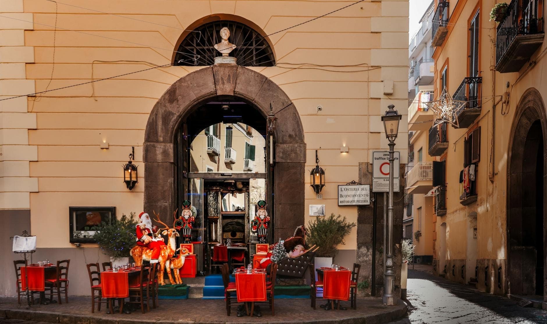 Rua encantadora de Nápoles com mesas ao ar livre, toalhas vermelhas, decorações natalinas e figuras típicas em frente a um prédio histórico.