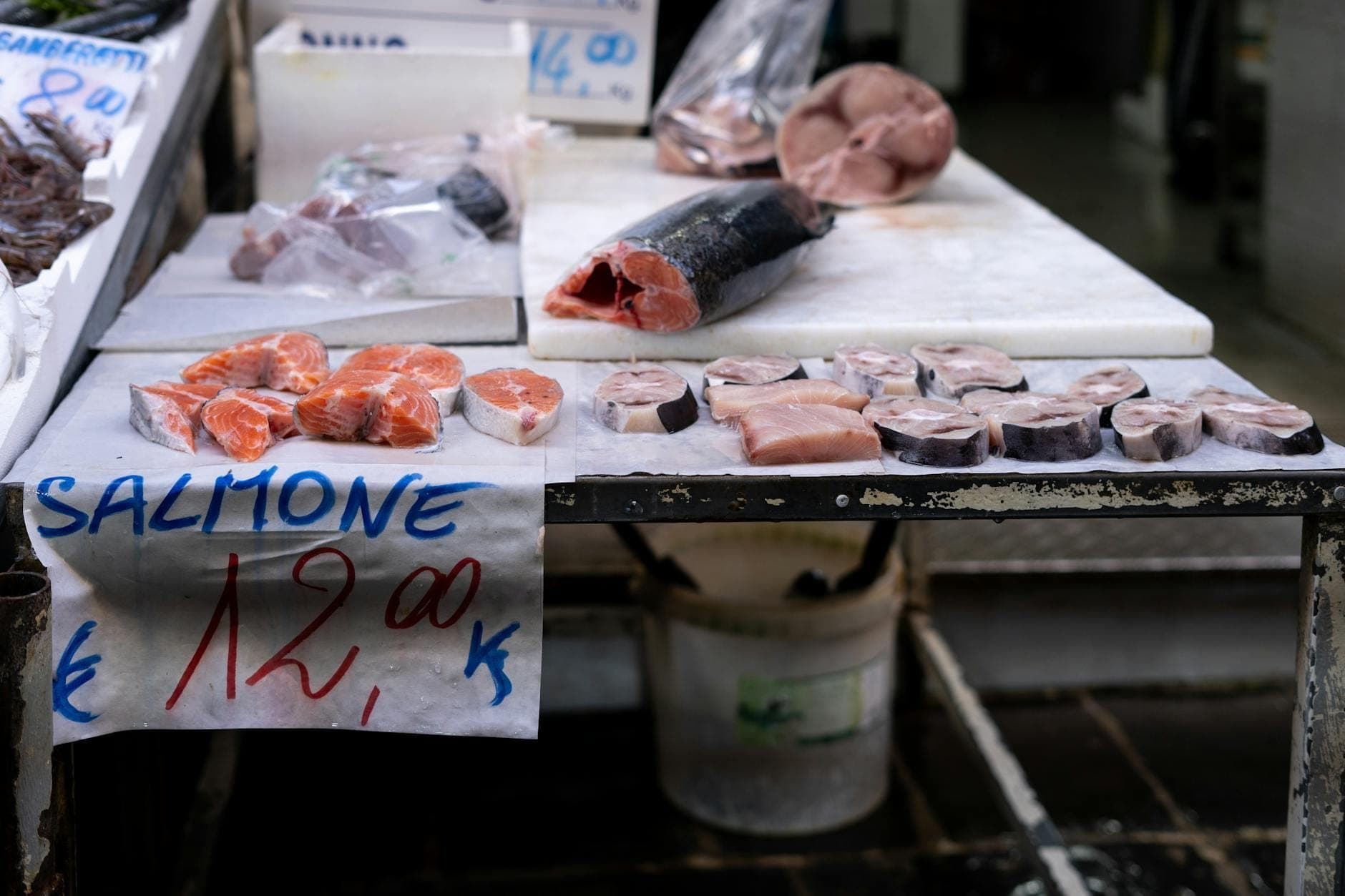 Imagem aproximada de um balcão de peixaria exibindo filés e fatias de salmão fresco, com placas de preço escritas à mão em italiano, representando os ingredientes tradicionais da cozinha napolitana.