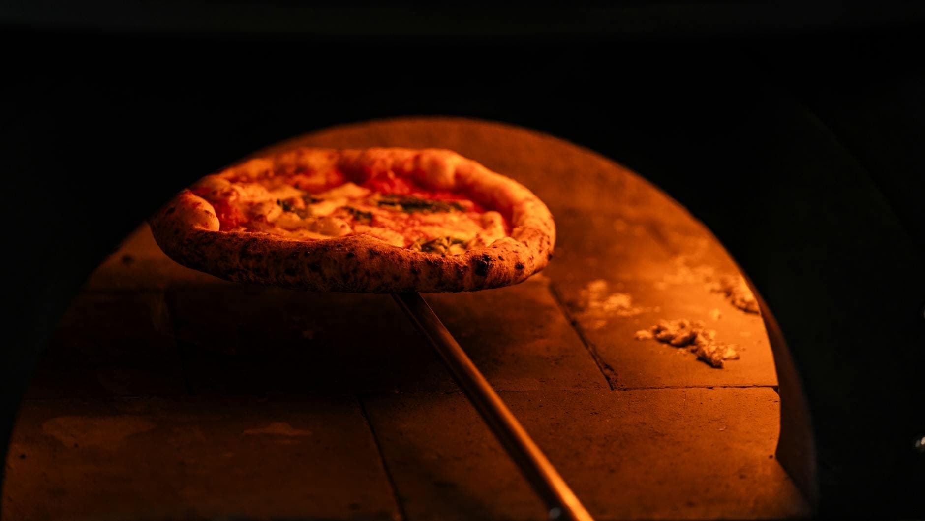 Vista de perto de uma pizza napolitana sendo assada em um forno a lenha, com chamas visíveis e borda chamuscada.