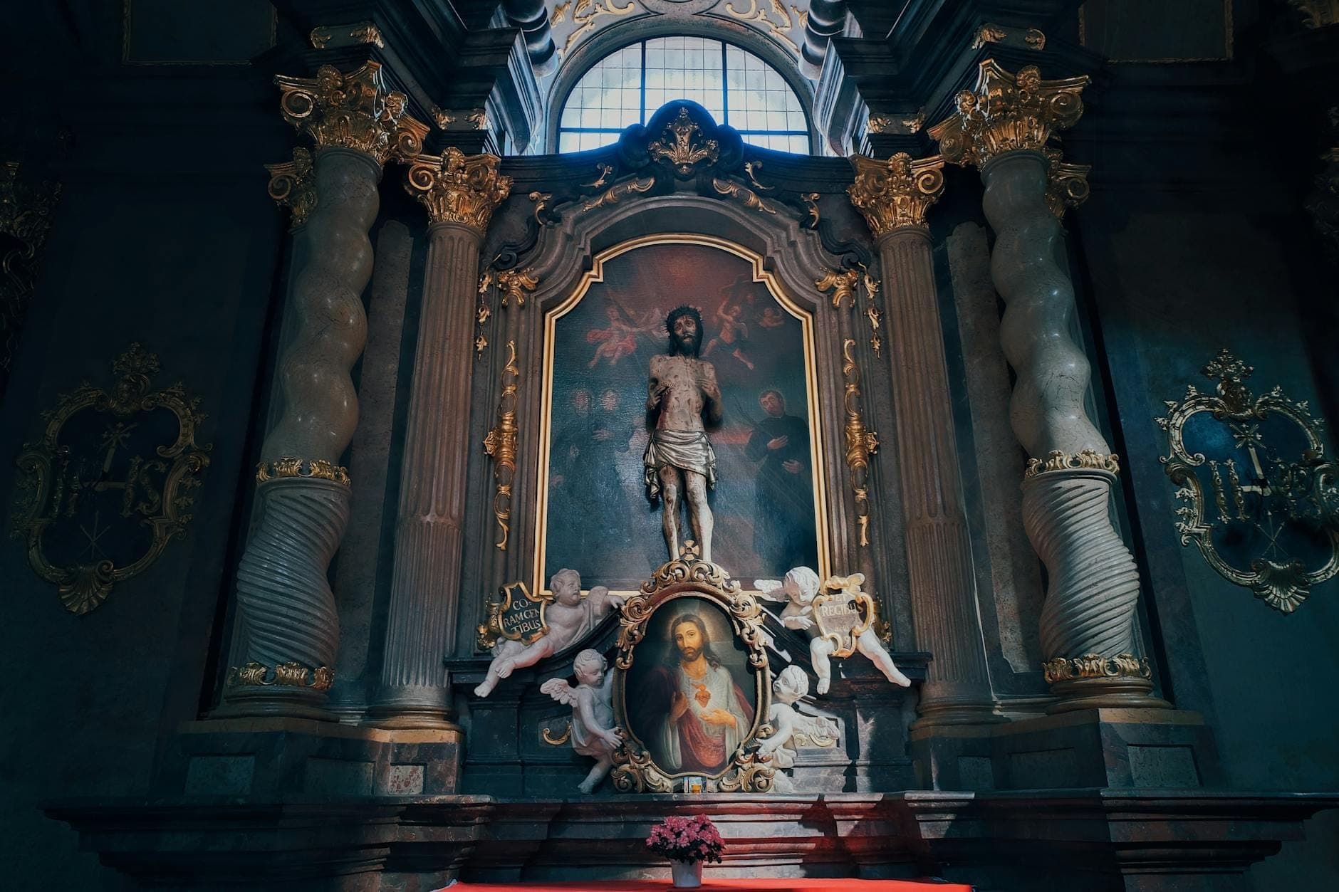 Interior de capela barroca ornamentada com uma escultura de Cristo e anjos, decorações douradas e uma grande janela arqueada acima.