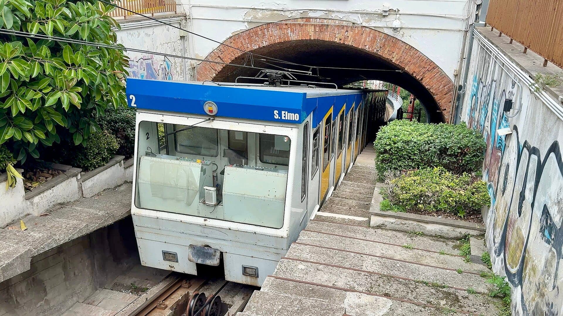 O vagão do funicular de Nápoles S. Elmo sai de um túnel de tijolos, ladeado por arbustos verdes e paredes com grafites sob a luz do dia nublada.
