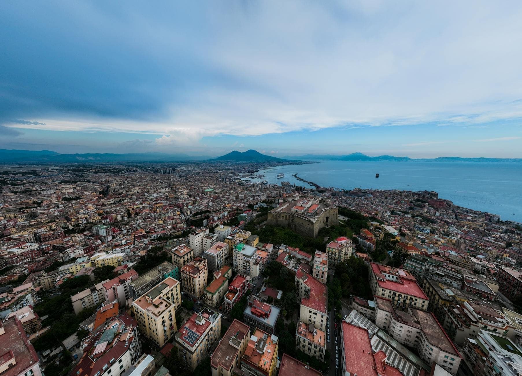 Vista aérea de Nápoles com o mar ao sul, o Monte Vesúvio ao fundo e os edifícios da cidade espalhados abaixo sob um céu azul.