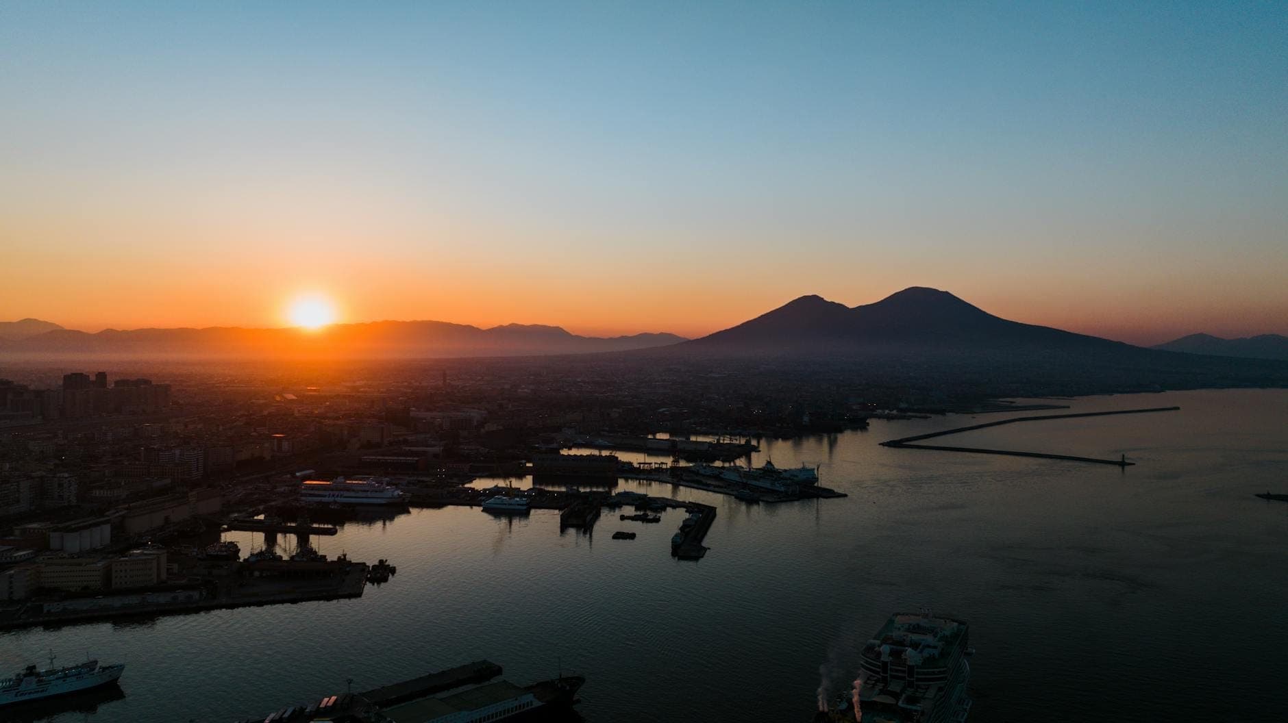 Vista aérea sobre o Golfo de Nápoles ao pôr do sol com o Monte Vesúvio ao fundo e edifícios da cidade ao longo da orla marítima.
