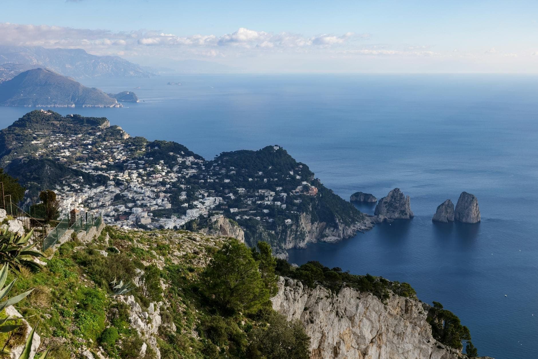 Vista panorâmica da ilha de Capri com edifícios caiados de branco, falésias dramáticas, os Faraglioni e o mar estendendo-se em direção à costa de Nápoles.