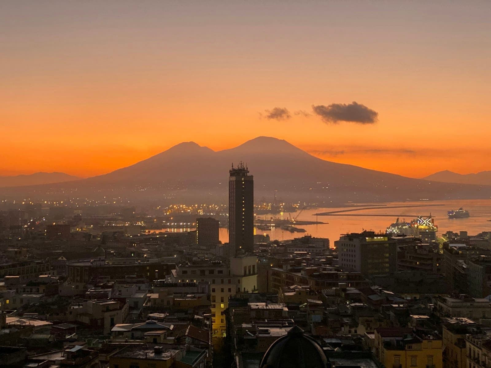 Vista panorâmica do pôr do sol sobre a paisagem urbana de Nápoles com o Monte Vesúvio, céu resplandecente, porto e edifícios iluminados pelas luzes da tarde.