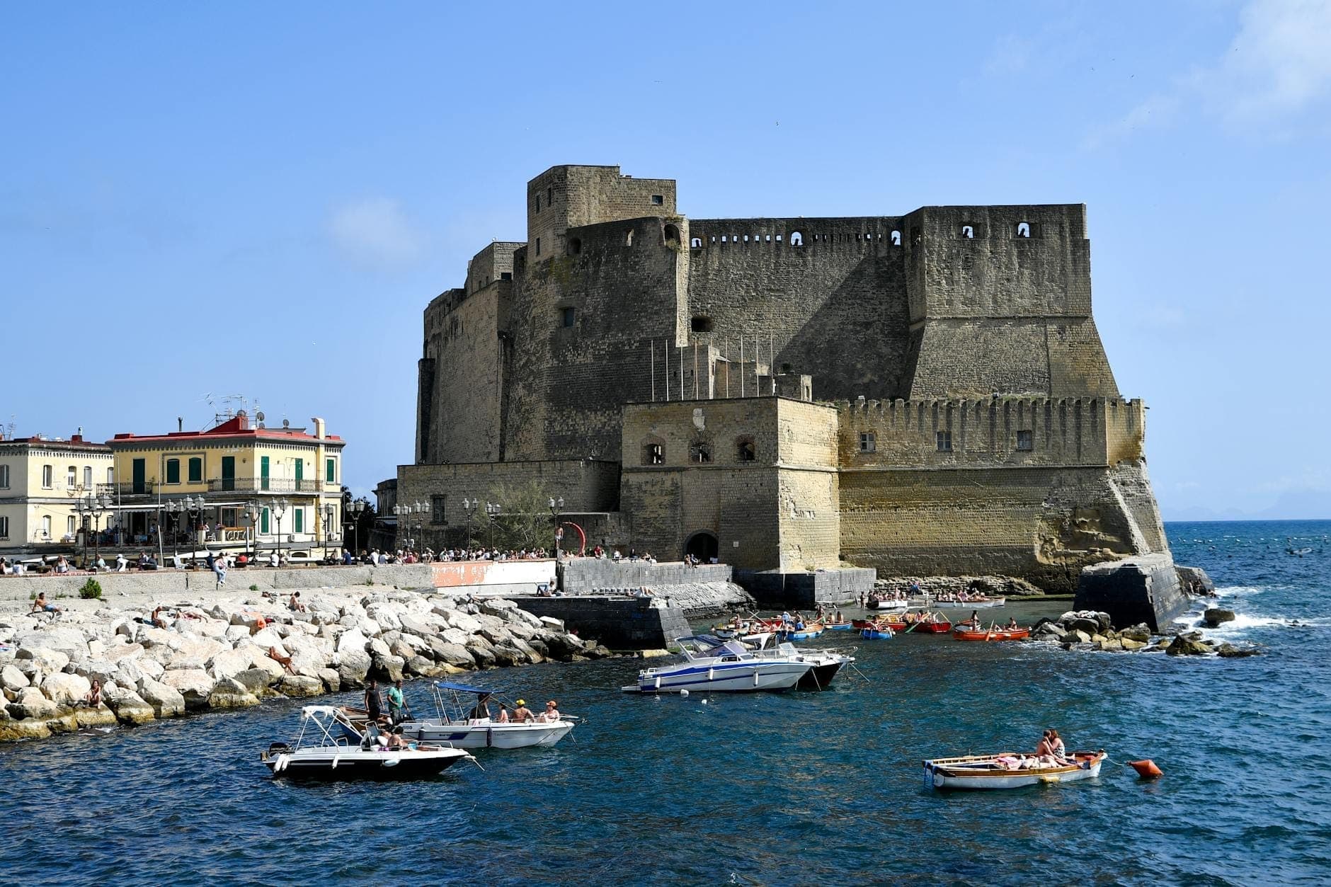Castel dell'Ovo na orla marítima de Nápoles com pequenos barcos em águas azuis e pessoas aproveitando a costa rochosa.