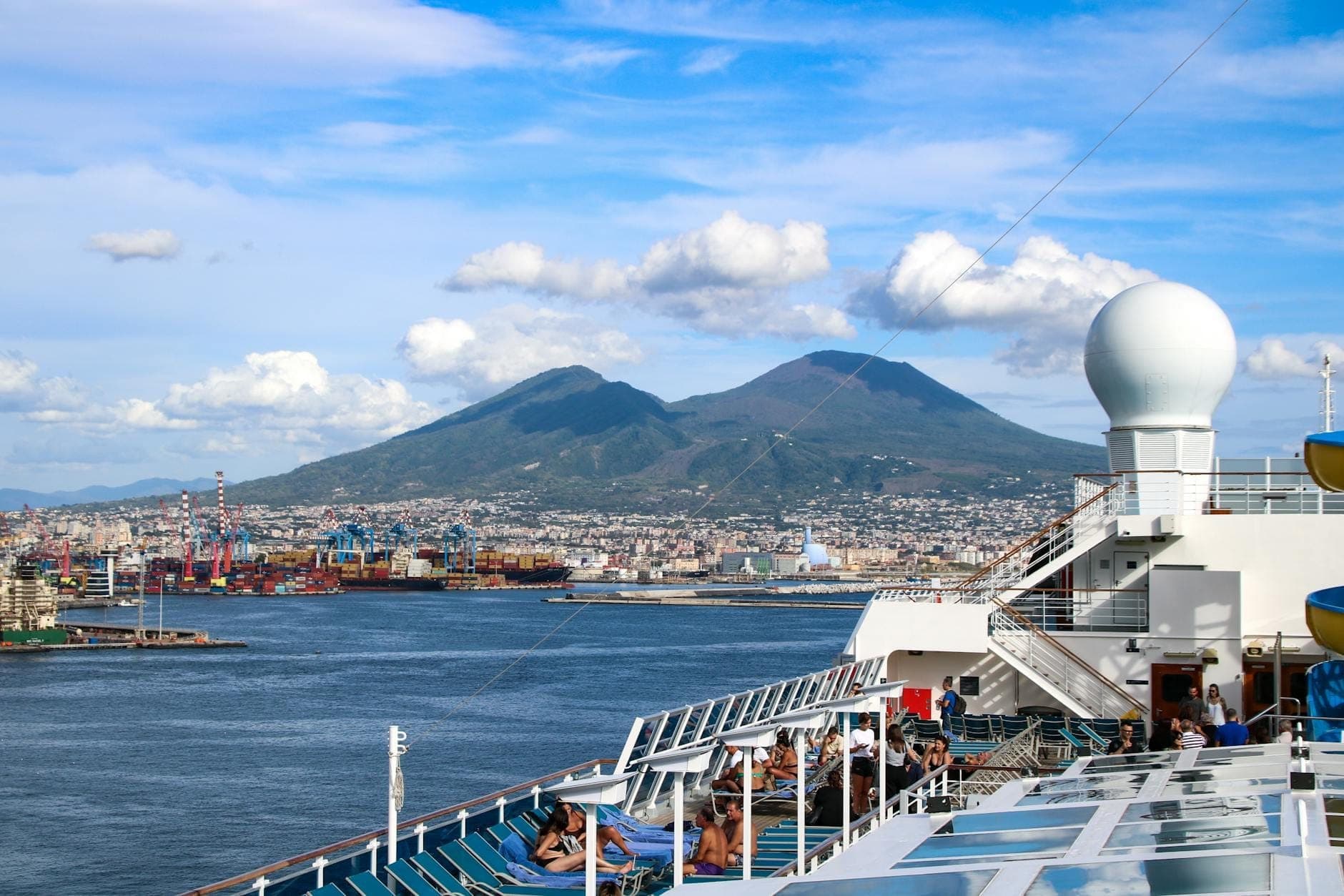 Deck de navio de cruzeiro com pessoas relaxando, com vista para a Baía de Nápoles, o porto da cidade e o Monte Vesúvio sob um céu azul brilhante.