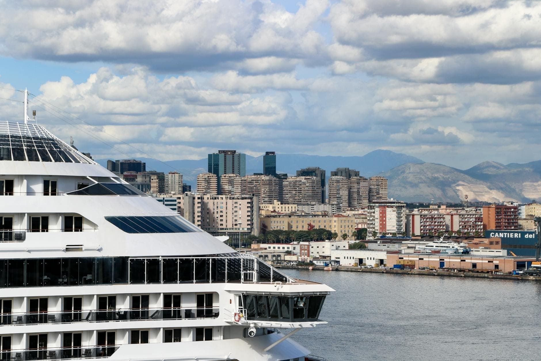 Navio de cruzeiro moderno atracado na orla marítima de Nápoles com edifícios altos, montanhas e céu nublado ao fundo.