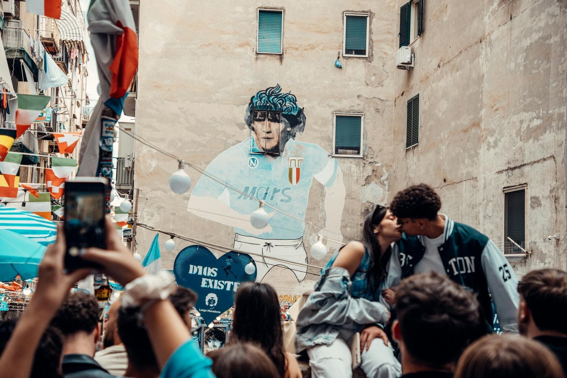 Uma rua movimentada de Nápoles com pessoas reunidas em torno de um grande mural de Maradona na parede de um prédio, decorado com bandeiras e enfeites coloridos.