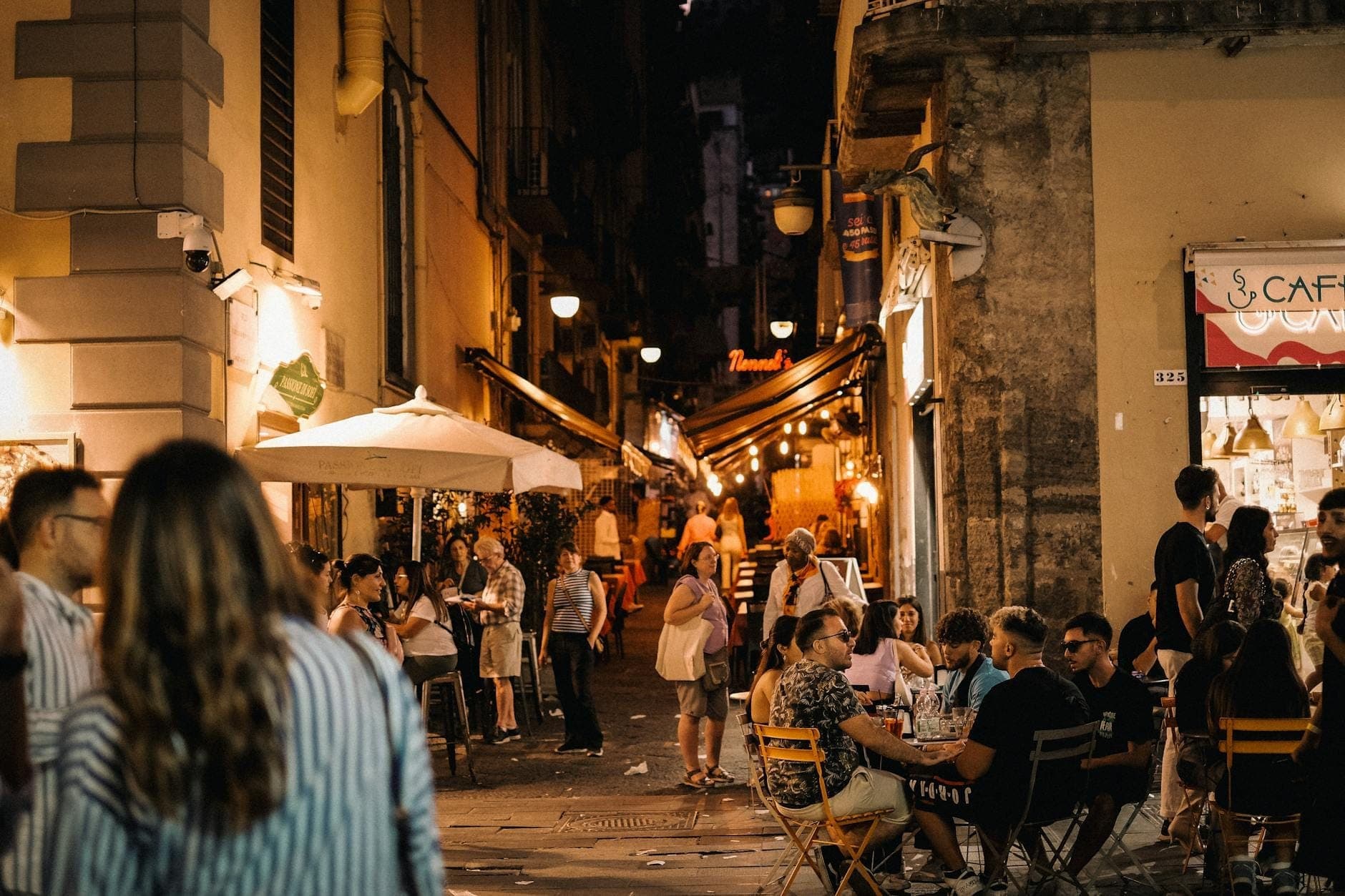 Movimentada rua de Nápoles à noite com pessoas jantando em cafés ao ar livre e caminhando por uma estreita viela iluminada.