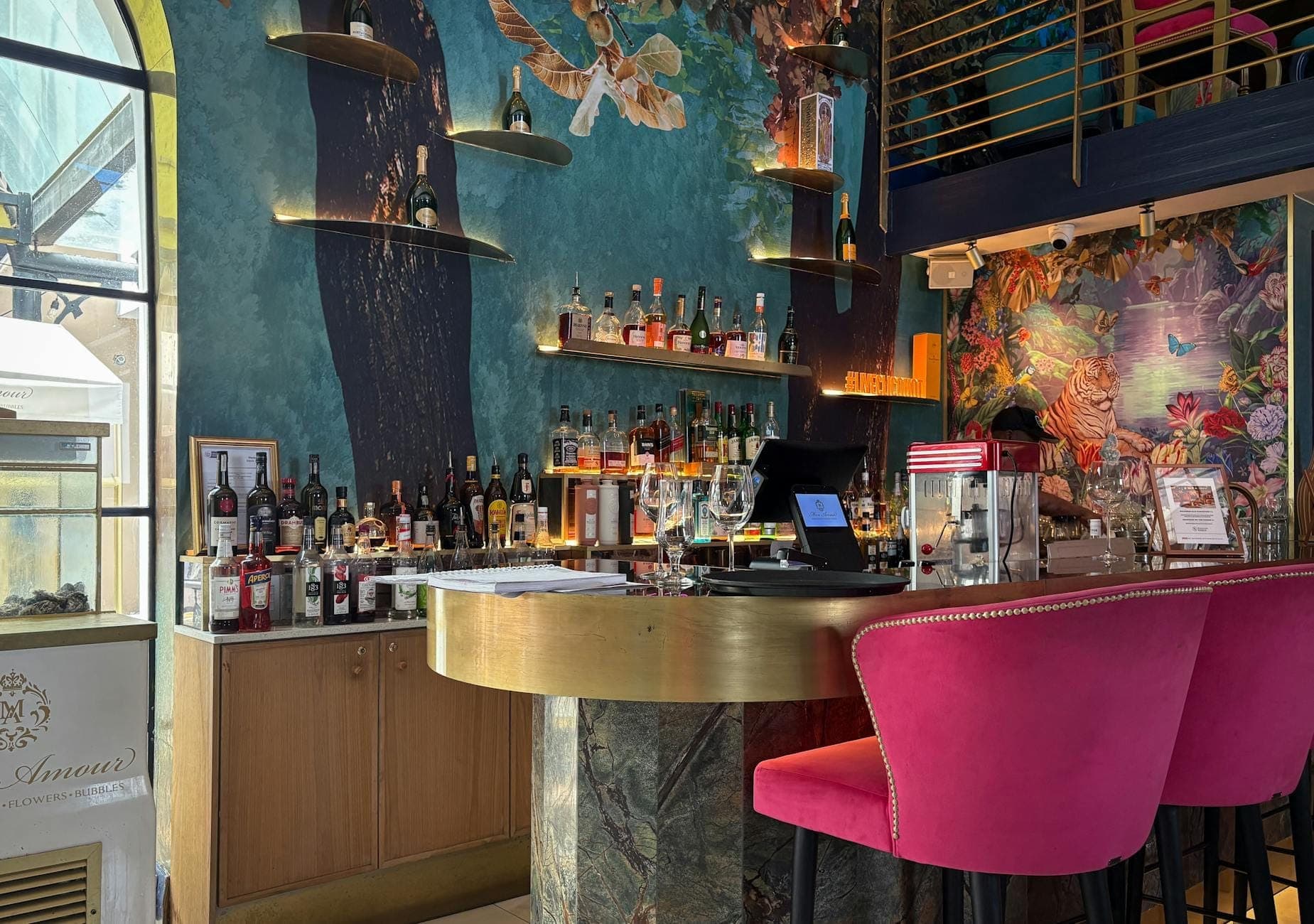 Bar de coquetéis chique e moderno em Nápoles com murais coloridos nas paredes, prateleiras de licores e confortáveis banquetas rosas em um ambiente animado e acolhedor.