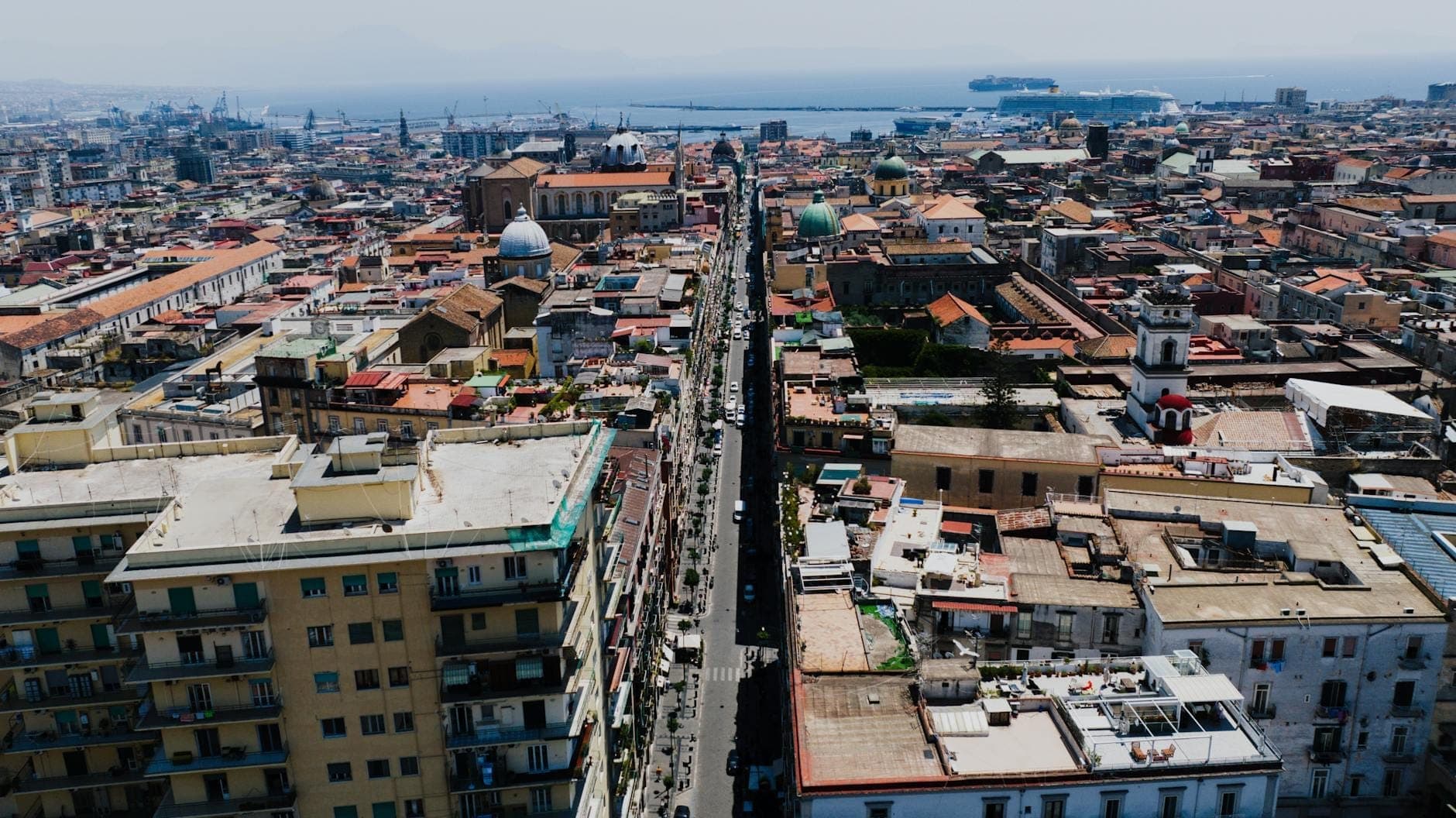 Vista aérea do centro histórico de Nápoles com uma longa rua reta em direção ao mar, cercada de edifícios antigos e cúpulas.