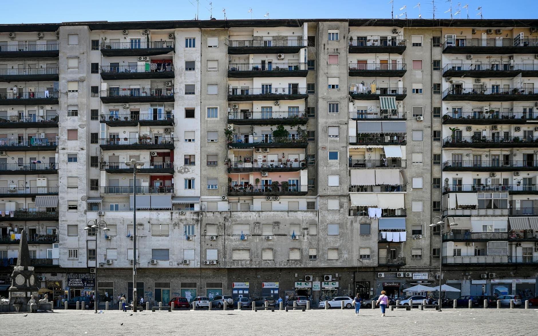 Fachada de um grande edifício de apartamentos desgastado em Nápoles com varandas visíveis, antenas parabólicas e roupa estendida, mostrando um caráter residencial acessível.