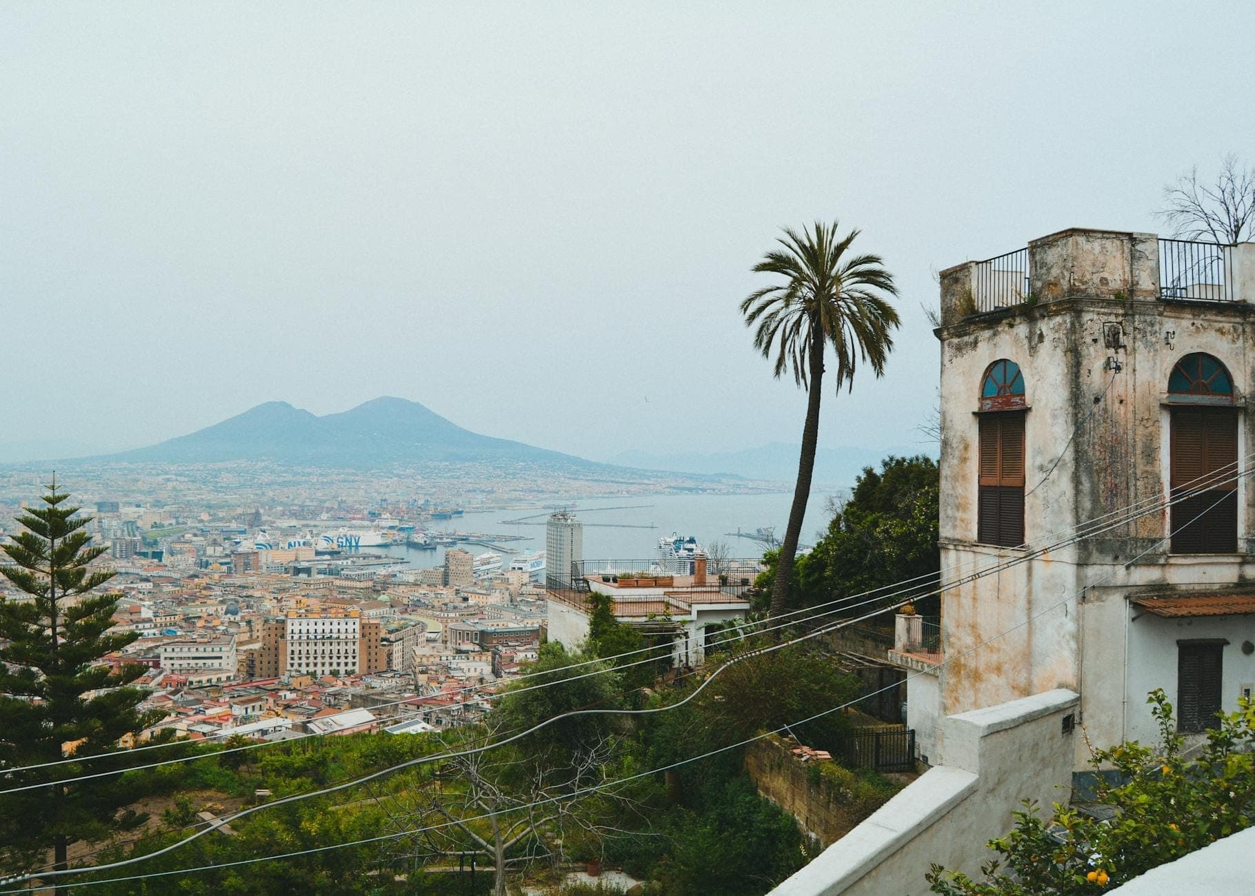 Vista sobre Nápoles com um edifício antigo, uma palmeira e o Monte Vesúvio ao fundo, captando a atmosfera da cidade e da baía.