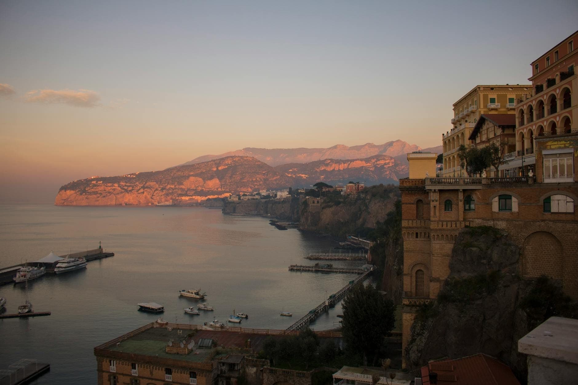 Vista dos imponentes penhascos de Sorrento e seus edifícios coloridos com vista para a calma Baía de Nápoles ao pôr do sol, com barcos ancorados abaixo.