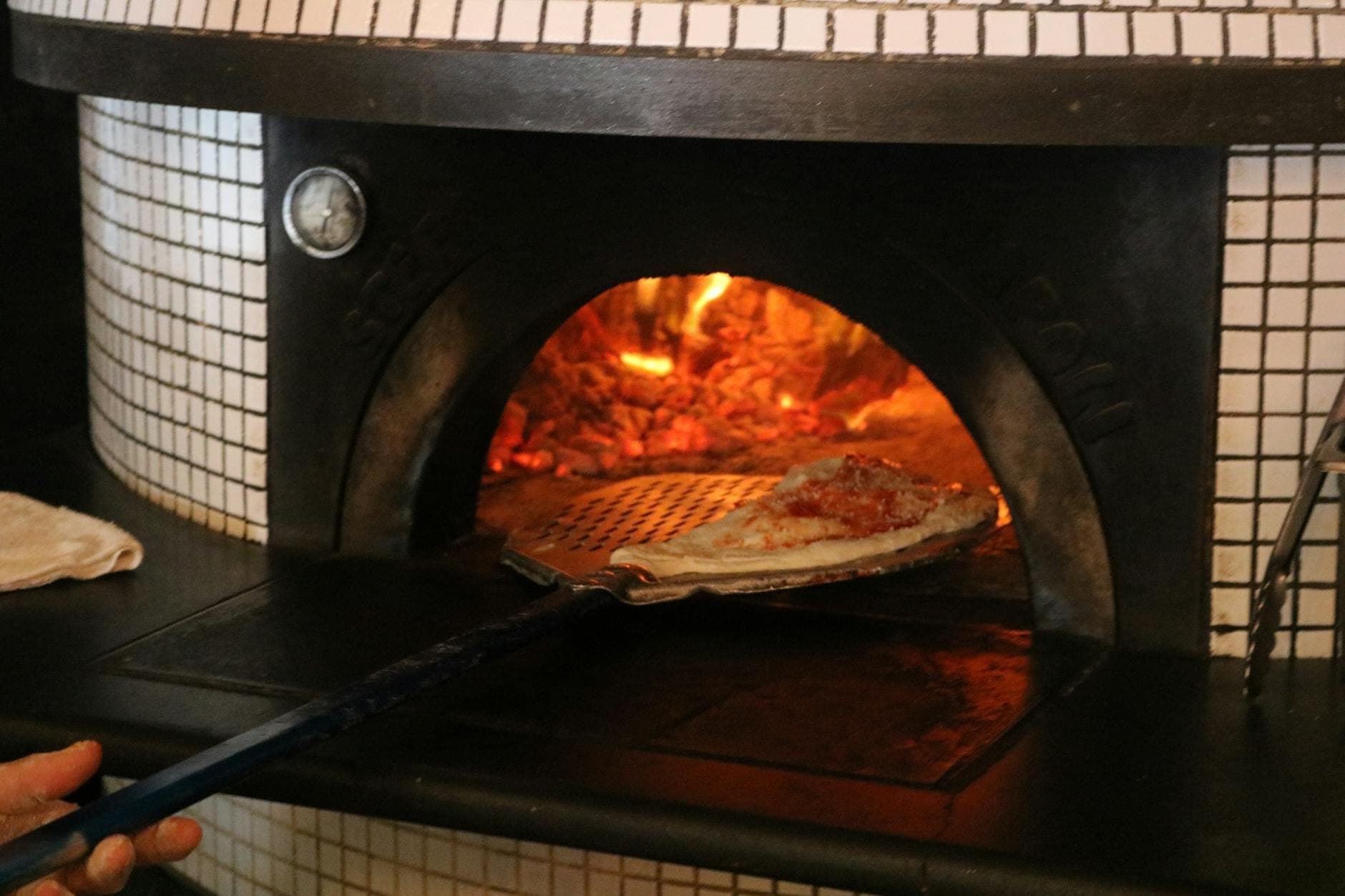 Pizza sendo colocada em um forno a lenha tradicional com brasas acesas, ilustrando a técnica de preparo da pizza napolitana.