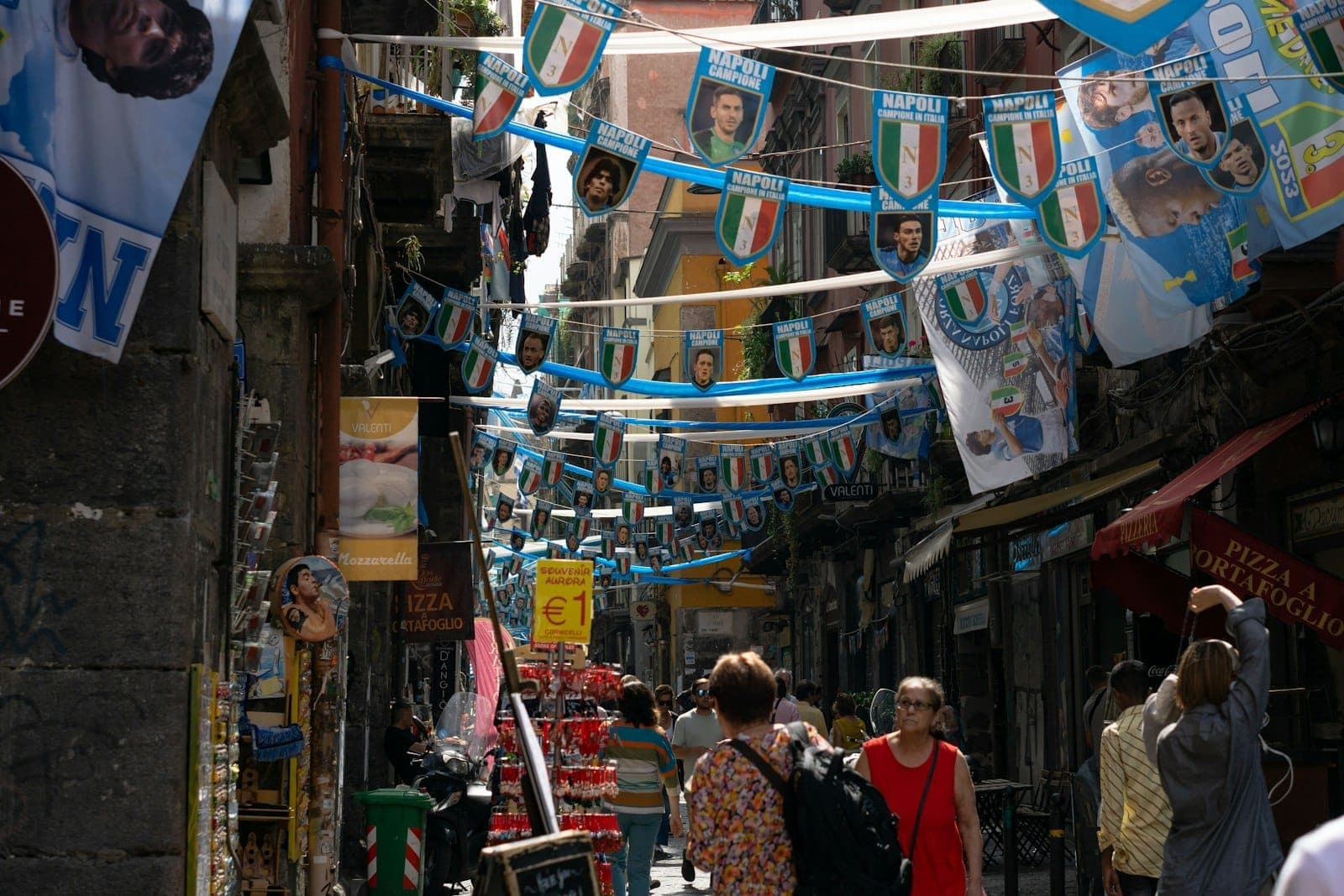 Uma animada cena de rua em Nápoles com bandeiras e faixas, pessoas caminhando e edifícios estreitos muito próximos entre si.