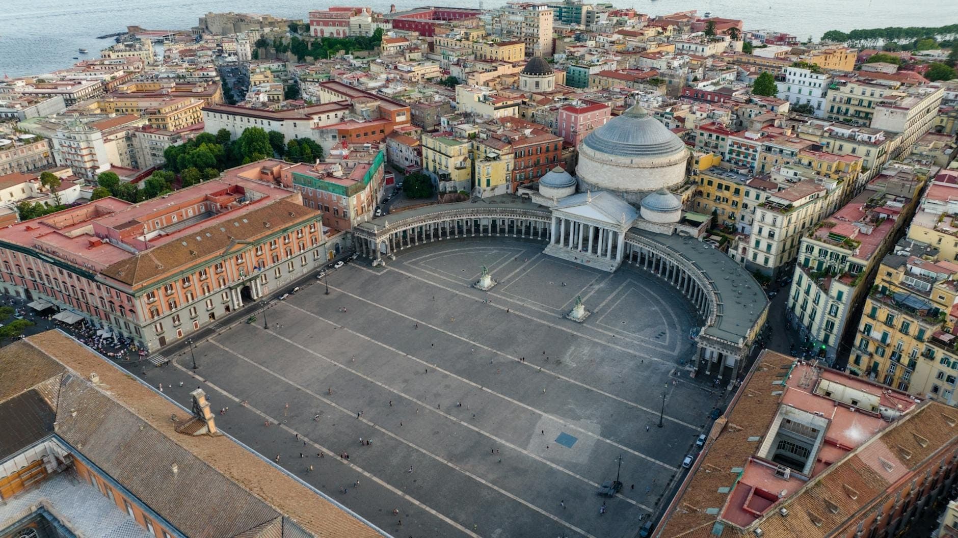 Vista aérea da Piazza del Plebiscito e de San Francesco di Paola com a paisagem urbana de Nápoles e a baía ao fundo.