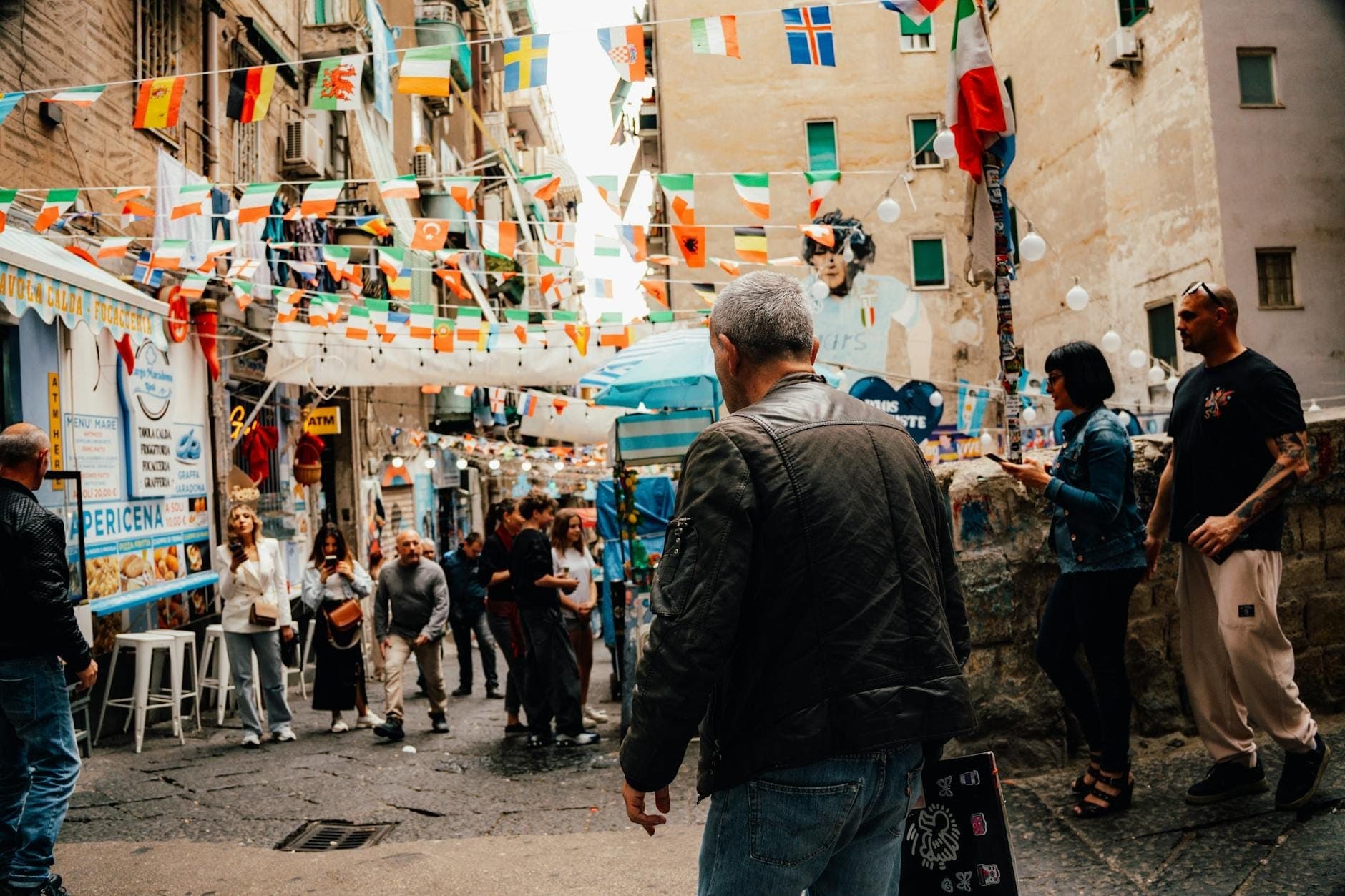 Movimentada rua de Nápoles decorada com bandeiras, pessoas caminhando e barracas de comida, animada atmosfera de festival.