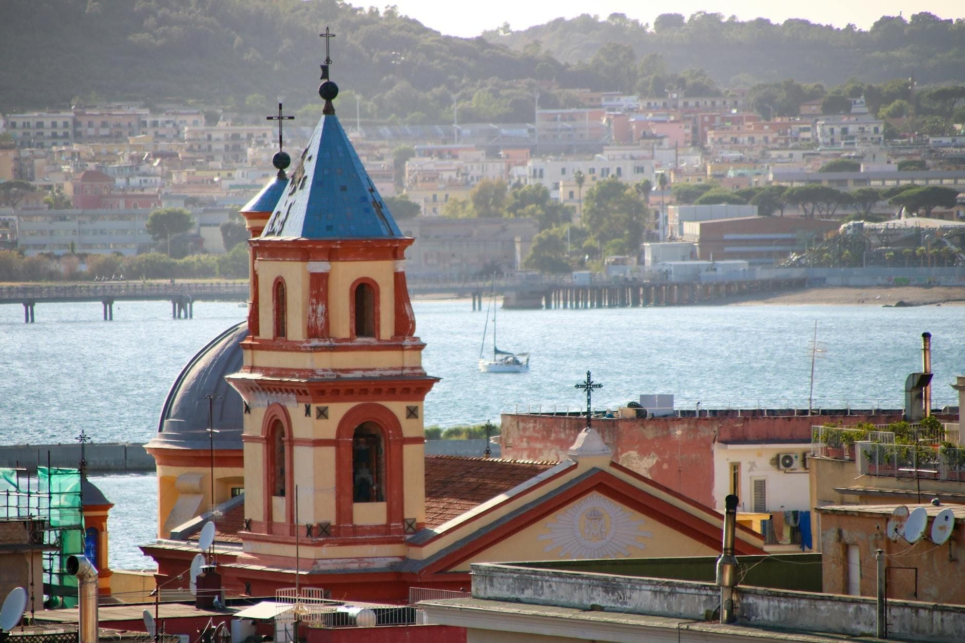 Torre de igreja com uma agulha azul e cruz com vista para o calçadão de Nápoles, com casas e água visíveis ao fundo.