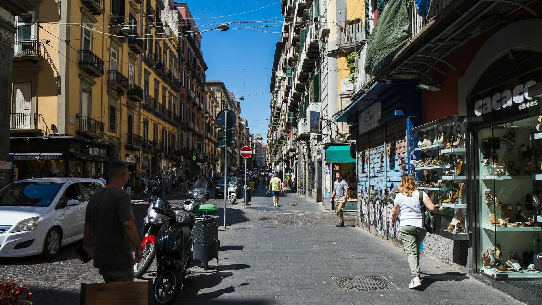 Rua comercial movimentada em Nápoles com pessoas caminhando, prédios coloridos, vitrines e uma atmosfera urbana animada sob um céu azul.