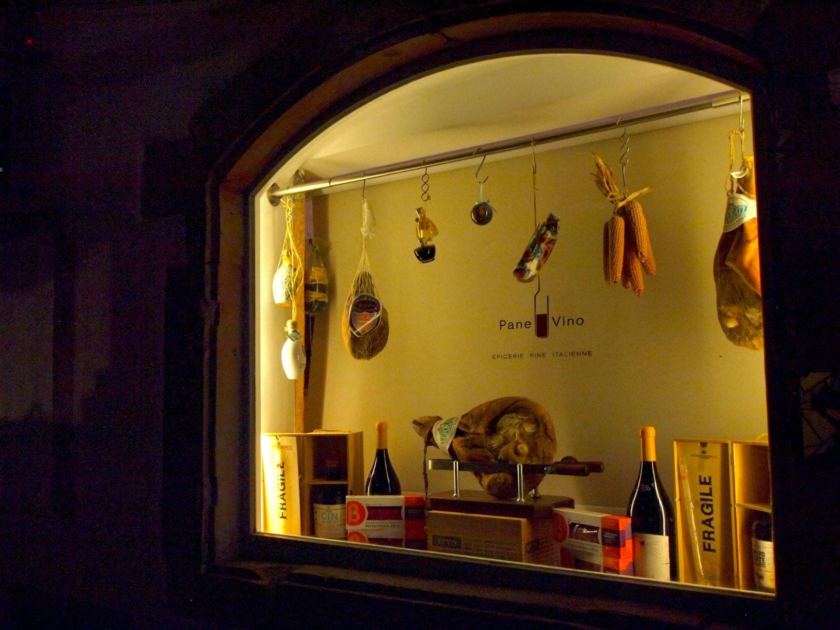 Vitrine de uma delicatessen com carnes curadas penduradas, garrafas de vinho, queijos e produtos alimentícios italianos, com iluminação aconchegante à noite.