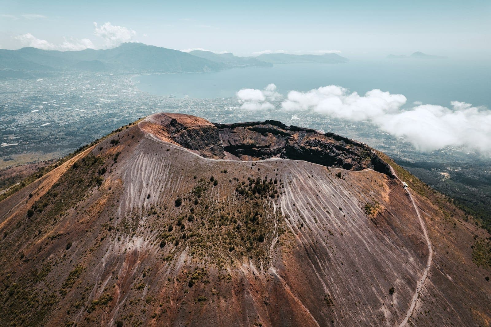 Vista aérea diretamente sobre a dramática cratera do Monte Vesúvio com a costeira Nápoles e o mar azul ao fundo.