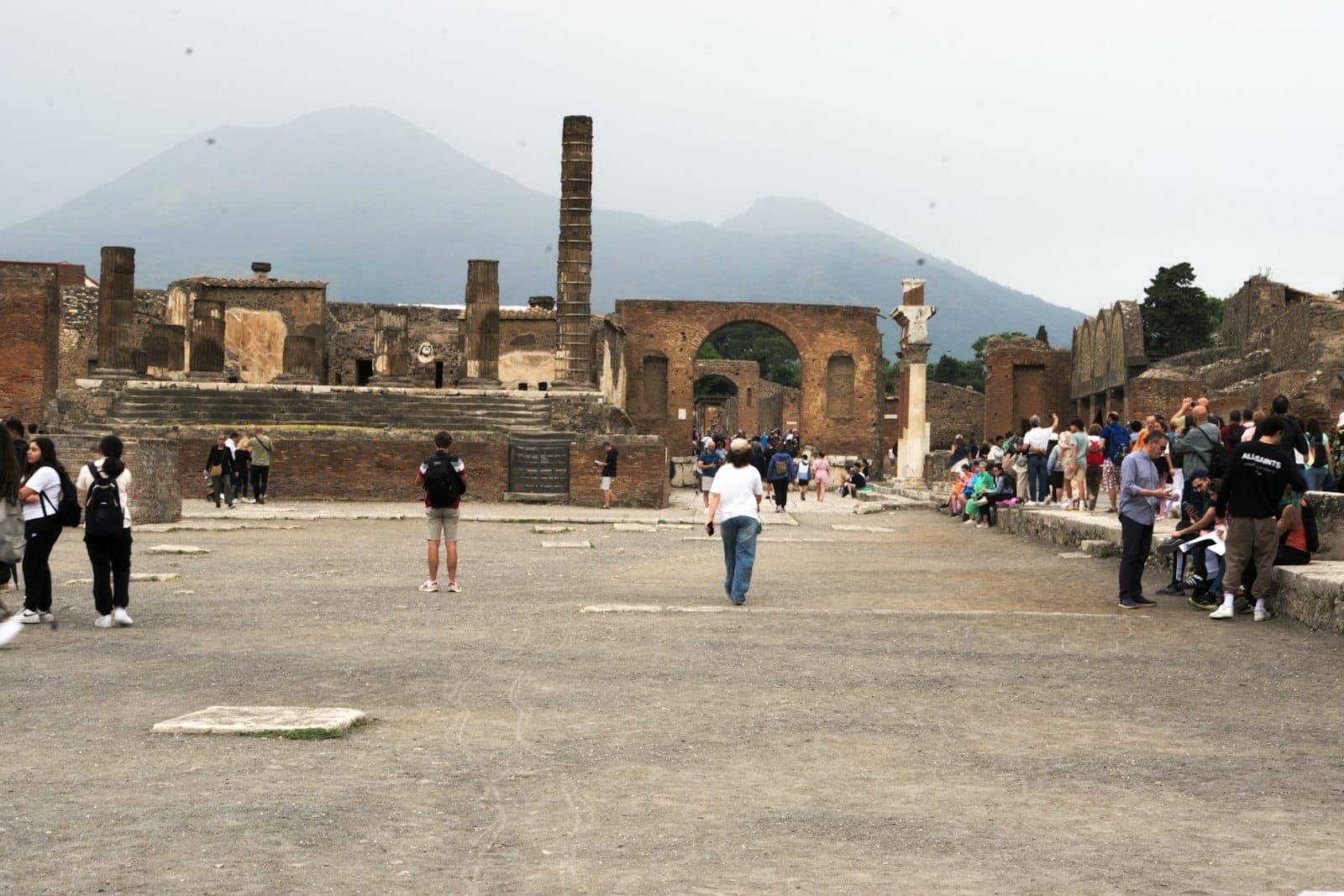 Turistas caminhando entre as ruínas de Pompeia com o Monte Vesúvio visível ao fundo, área do fórum central.