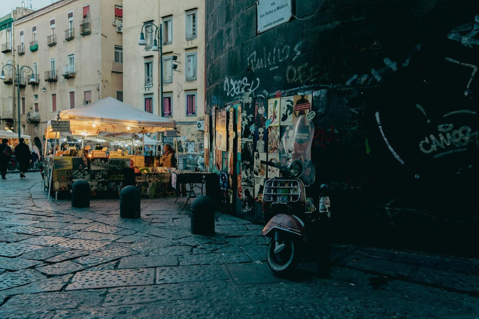 Cena noturna de uma rua em Nápoles com uma barraca de comida sob uma cobertura ao lado de edifícios desgastados da cidade e uma scooter estacionada.