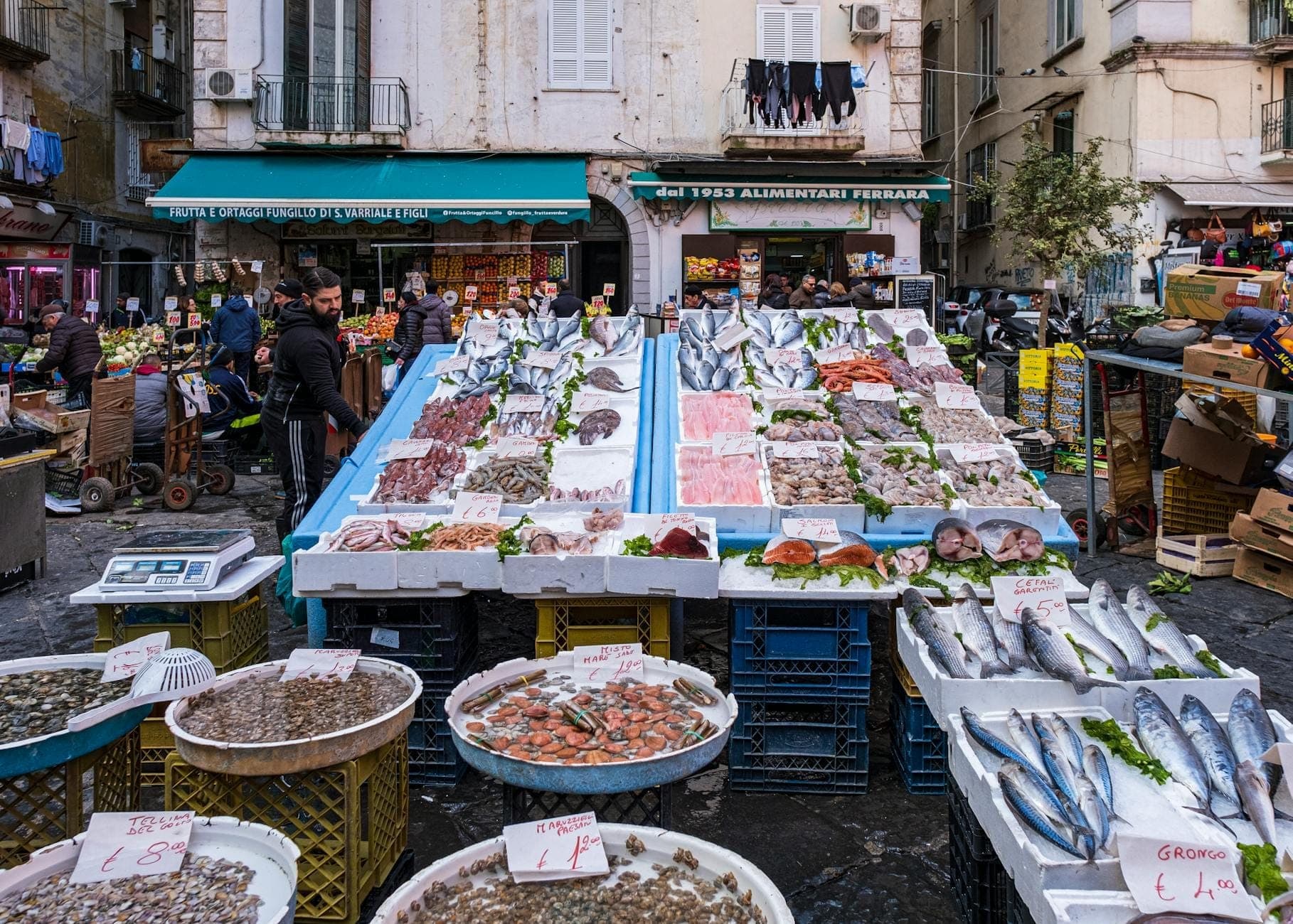 Cena de mercado ao ar livre em Nápoles com abundante marisco fresco exposto em mesas e pessoas locais fazendo compras em uma movimentada rua da cidade.