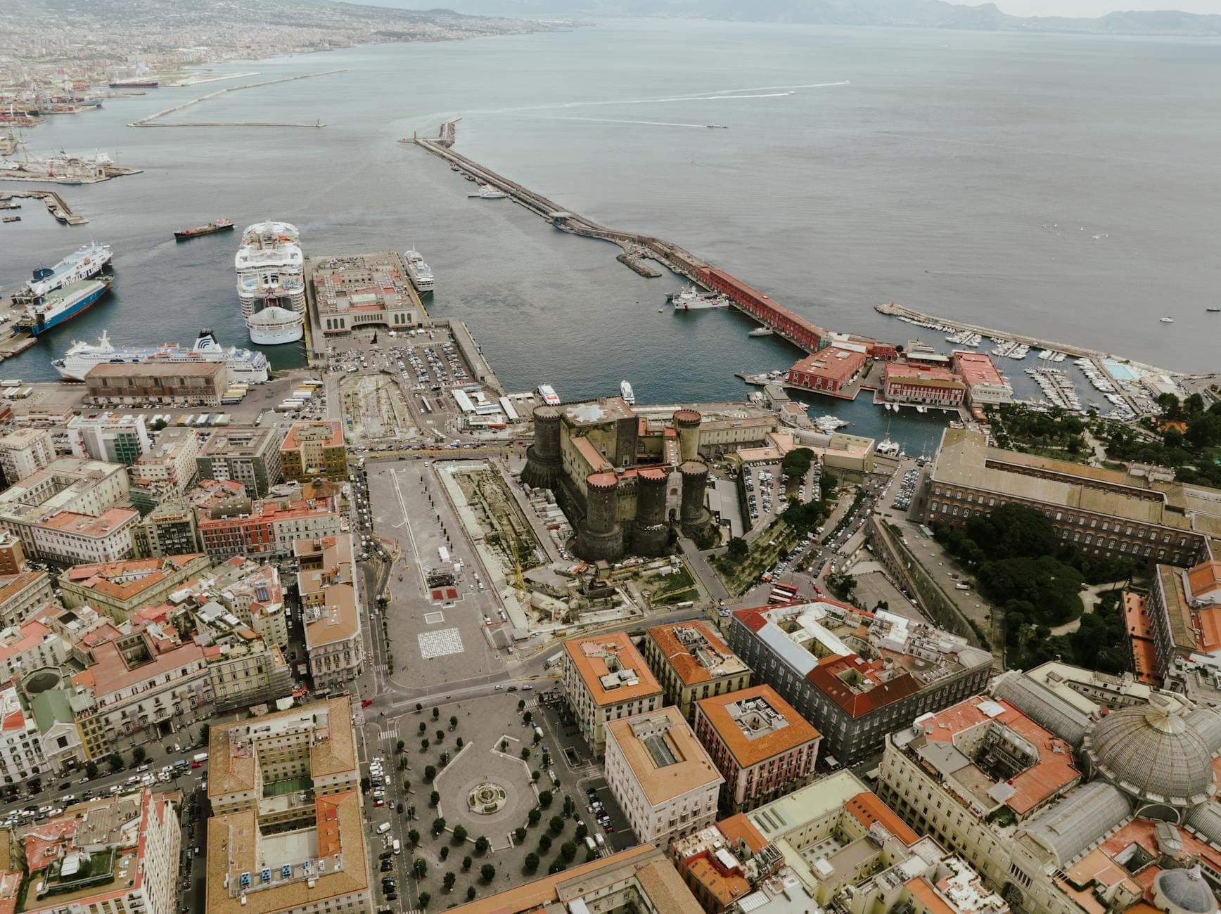 Vista aérea do porto de Nápoles com ferries, navios de cruzeiro, infraestrutura portuária e o Castello Maschio Angioino, mostrando as principais opções de transporte da cidade.