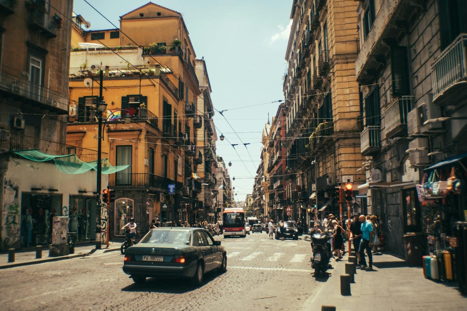 Rua movimentada de Nápoles com carros, pessoas e edifícios iluminados pelo sol, ilustrando a superlotação urbana e o agito sazonal.