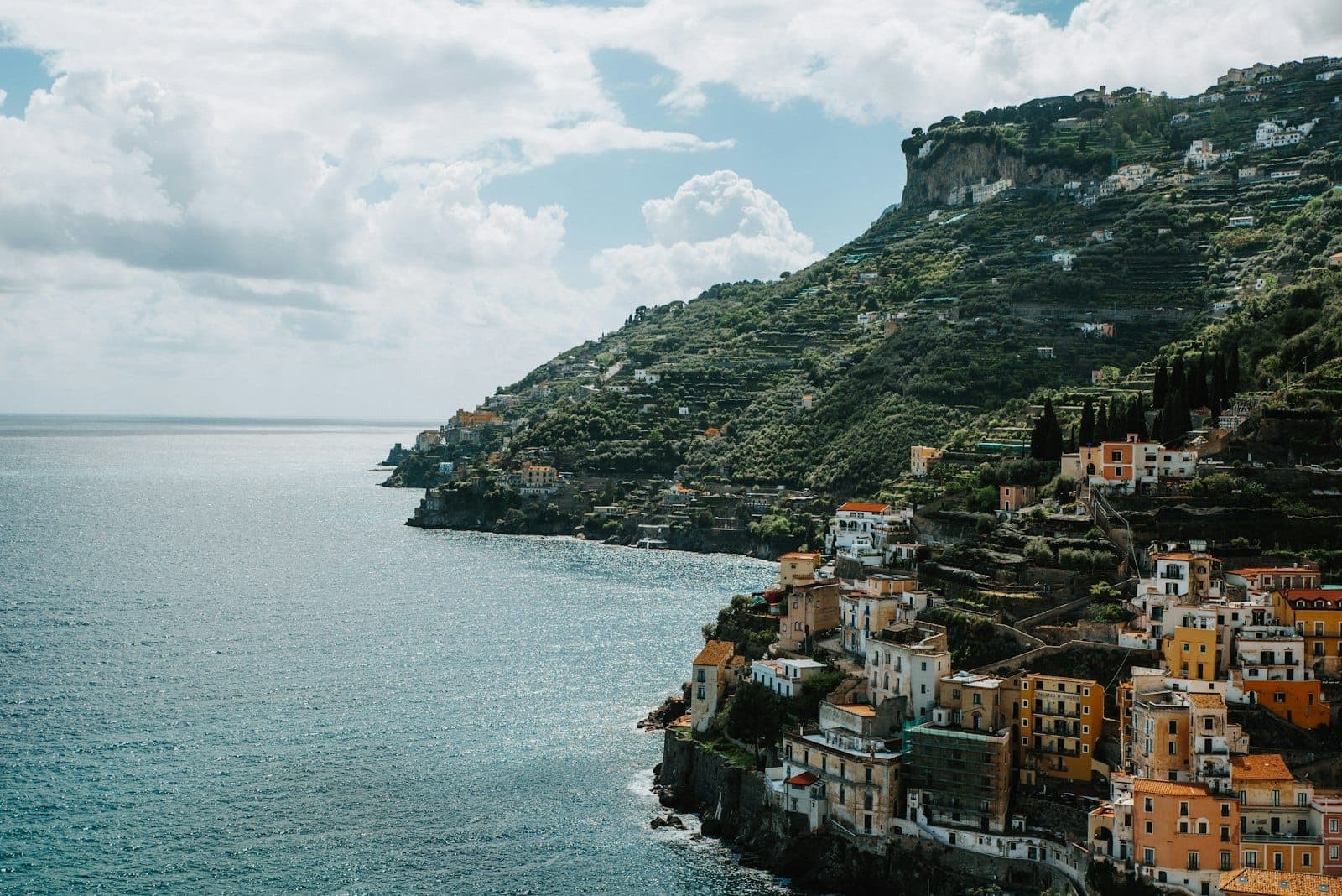 Costa dramática da Costa Amalfitana com casas coloridas nas falésias sobre o mar azul profundo e colinas verdes em terraços sob um céu parcialmente nublado.