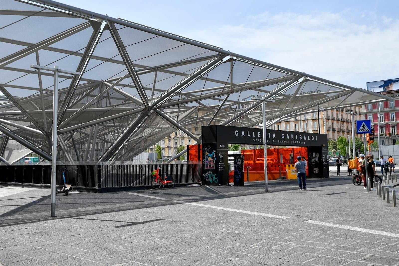 Entrada moderna da estação de trem Napoli Centrale com pessoas caminhando do lado de fora sob uma estrutura de teto de vidro geométrico.
