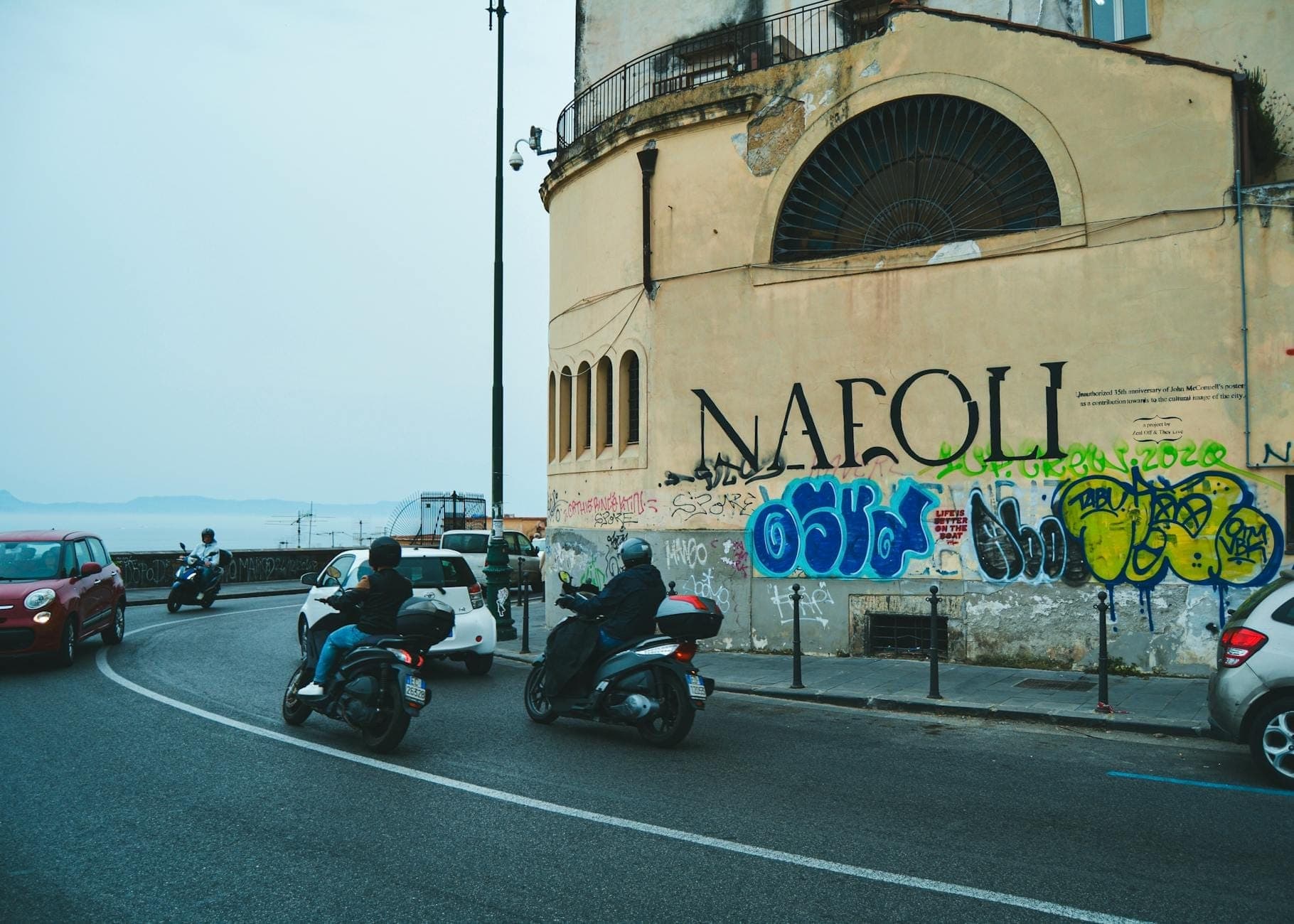 Scooters e carros trafegando por uma rua de Nápoles com grafites, captando o trânsito local e a atmosfera urbana.