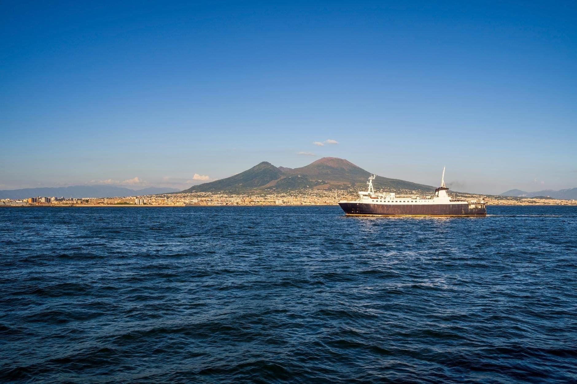 Uma balsa atravessando a Baía de Nápoles com o Monte Vesúvio e a cidade visíveis ao fundo em um dia claro.