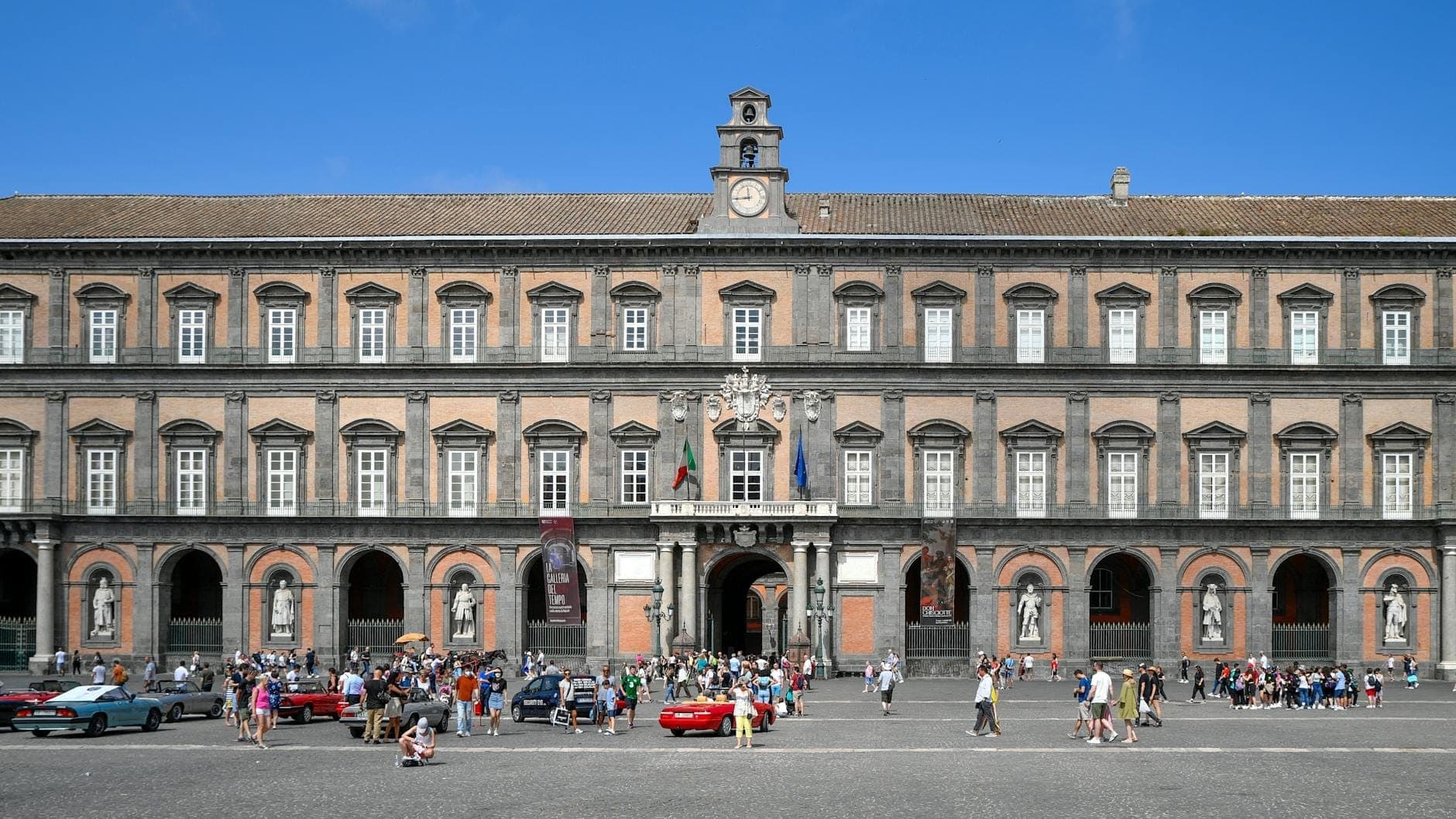 Grande fachada do Palazzo Reale di Napoli com pessoas caminhando e estátuas clássicas na praça.
