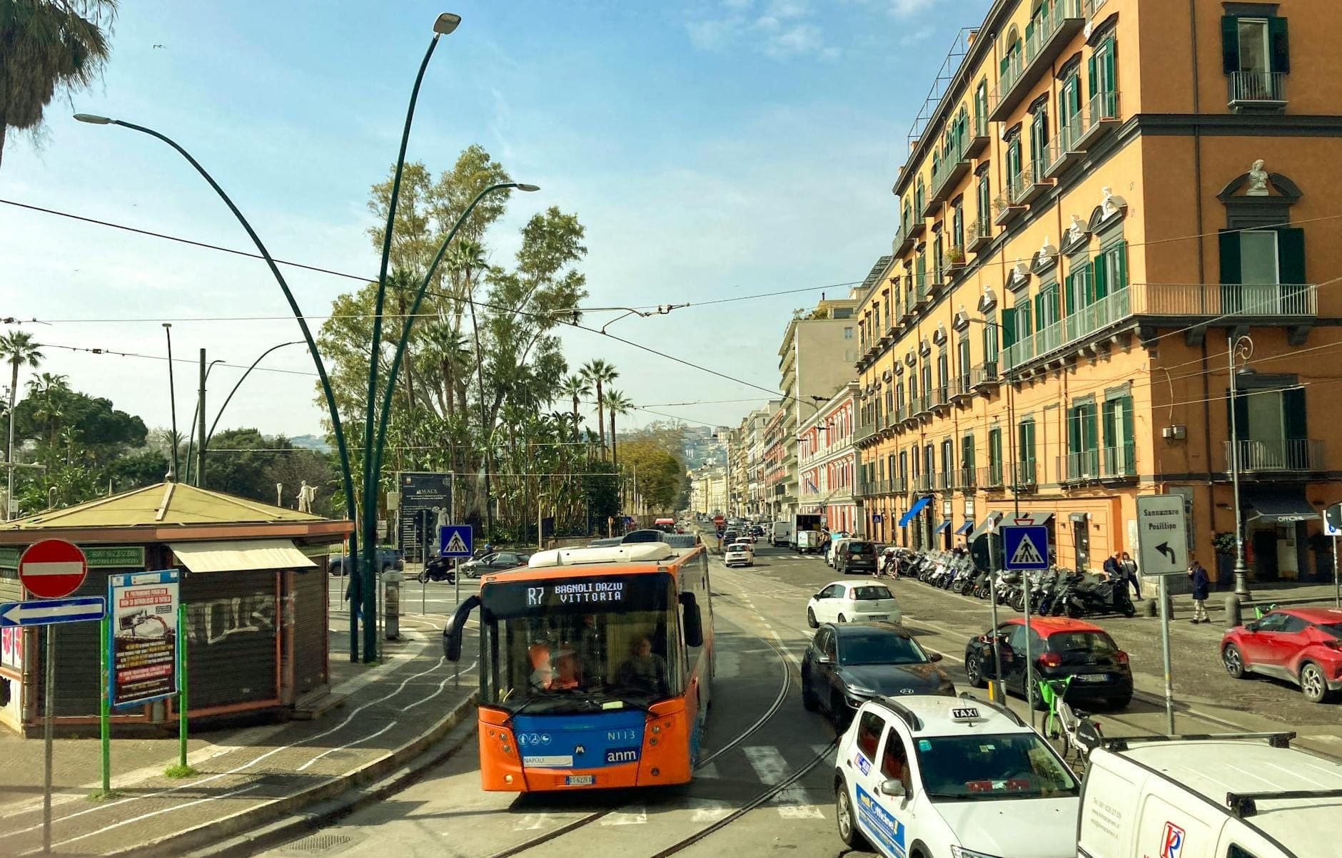 Ônibus urbano laranja, carros e táxis em uma movimentada rua de Nápoles com edifícios clássicos e um ponto de ônibus durante o dia.